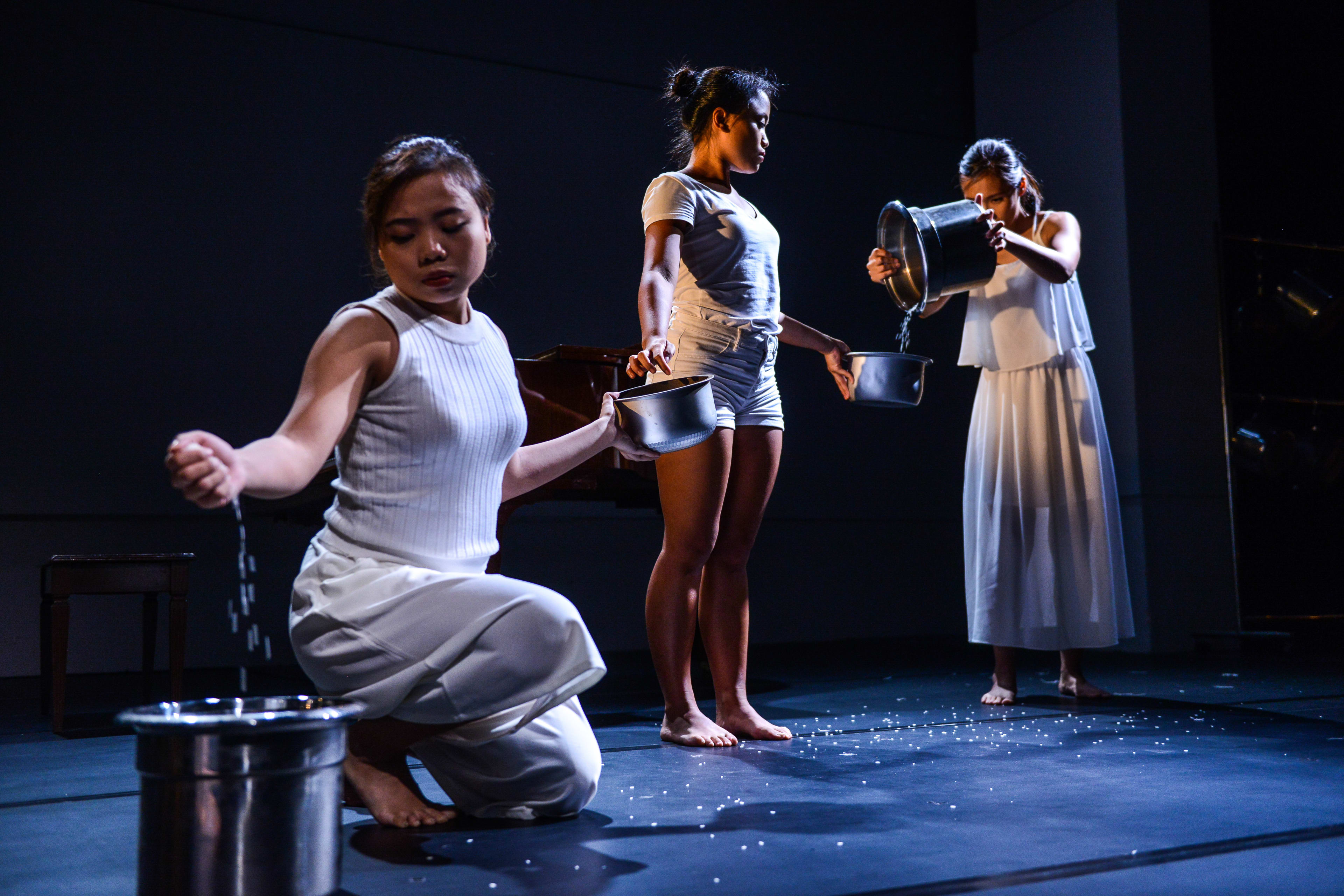 三位表演者身穿純白服飾，站立在天台製作（Rooftop Productions）當代劇場作品《不是女僕》(Not The Maids) 的極簡黑色舞台上。前景的表演者跪地，將手中的顆粒緩緩灑入金屬桶中，後方的兩位表演者則站立著，將盆中的顆粒傾倒而下。戲劇性的高對比燈光突顯了這種重複且具有儀式感的動作，成為該劇探討家務勞動議題的強烈視覺隱喻。此場景呈現了「全球化親職外判鏈」。