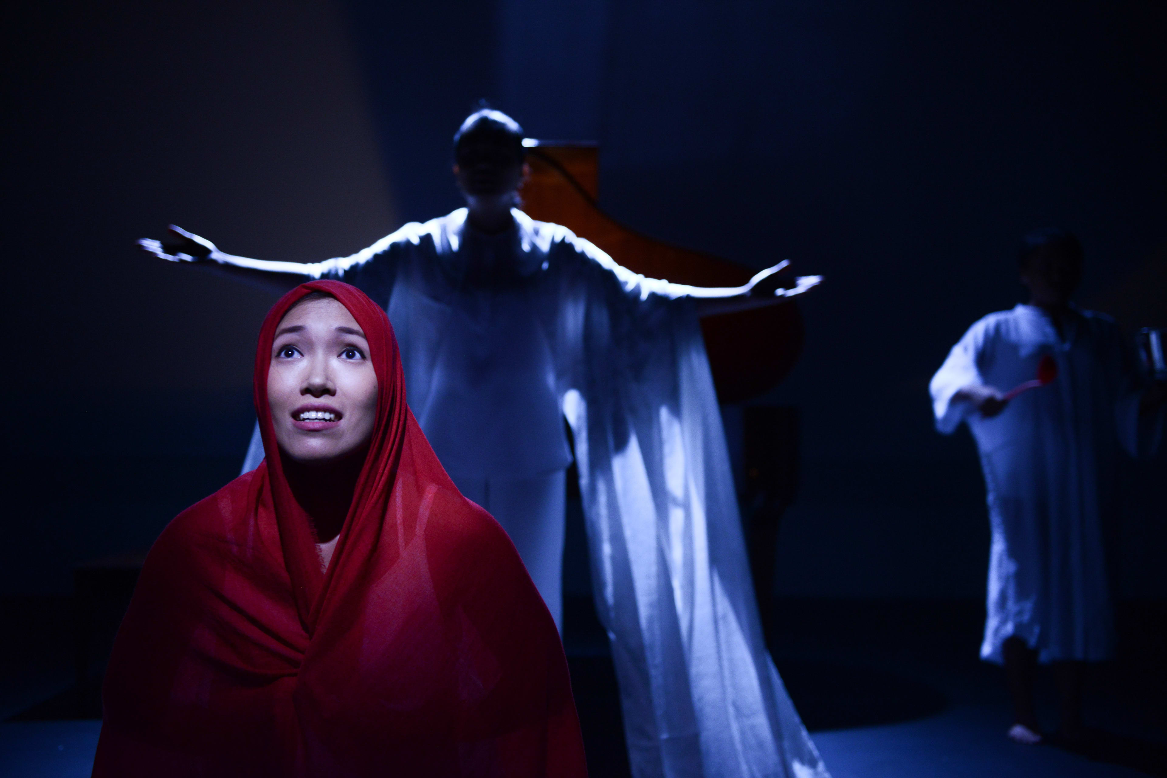 在天台製作 (Rooftop Productions) 的當代劇場作品《不是女僕》(Not The Maids) 中，演員梁皓貽（Isabella Leung）身披紅色頭巾位於畫面中央，表情中流露著深沉的渴望與脆弱。背景中，另一位表演者在冷冽的藍色舞台燈光下，雙臂向兩側伸展，呈現出莊嚴且具儀式感的姿態，右側則有另一名手持物件的表演者。極簡的舞美設計深刻體現了這部香港當代劇場作品的情感張力。表演者訴說著劇中的核心提問：「為什麼外傭必須要留宿於僱主家中？」