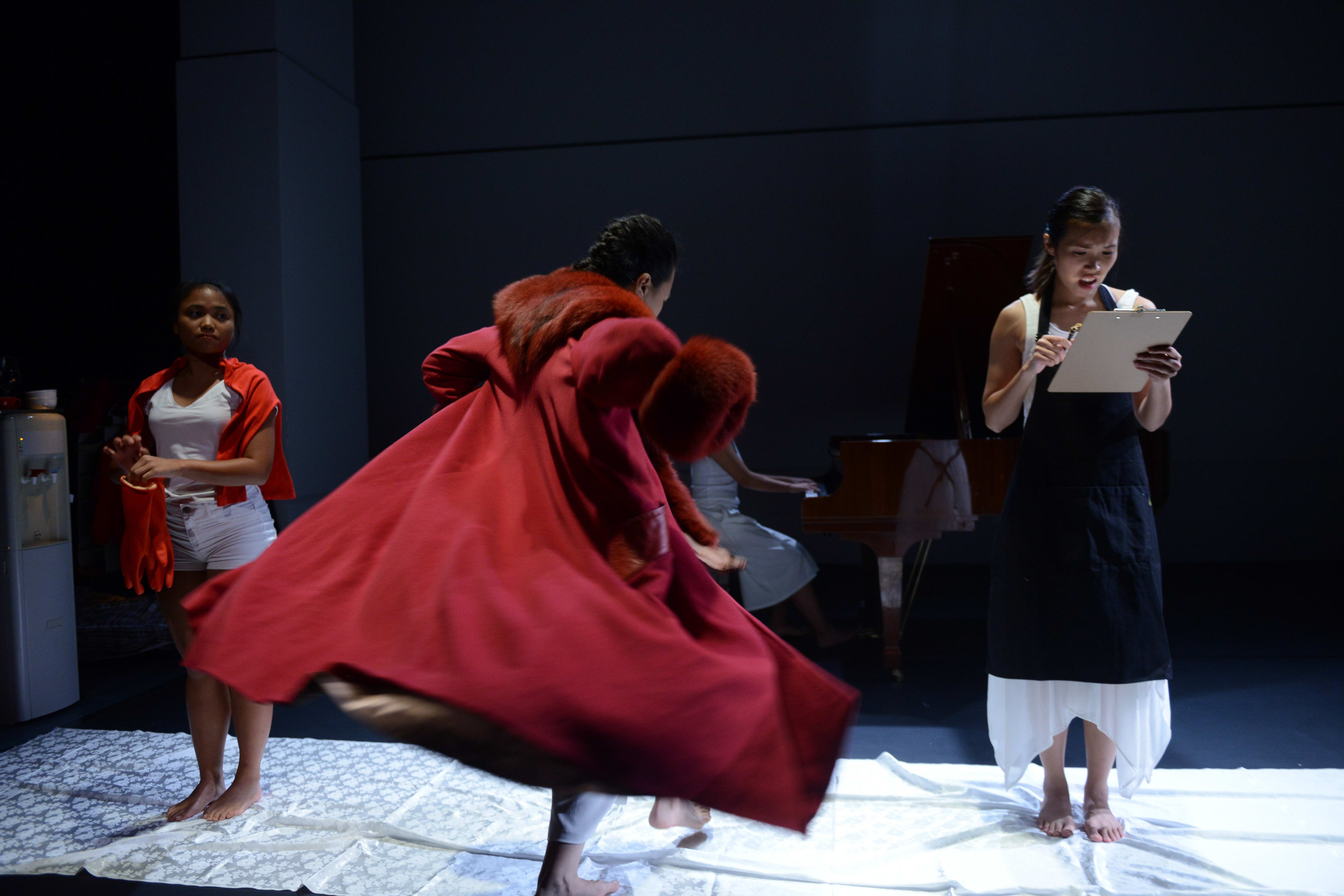 在天台製作 (Rooftop Productions) 的當代劇場作品《不是女僕》(Not The Maids) 中，一名演員身穿戲劇性的紅色大衣在舞台中央旋轉，梁皓貽 (Isabella Leung) 站在右側手持寫字板。左側是區洛思 (Russell Terre Aranza)，後方則可見一名鋼琴師在昏暗極簡的舞台佈景中伴奏。隨著演員進入角色情境，現場響起台詞：「成為監犯的人，會被帶進模擬監獄並須經過一系列的入獄程序。」