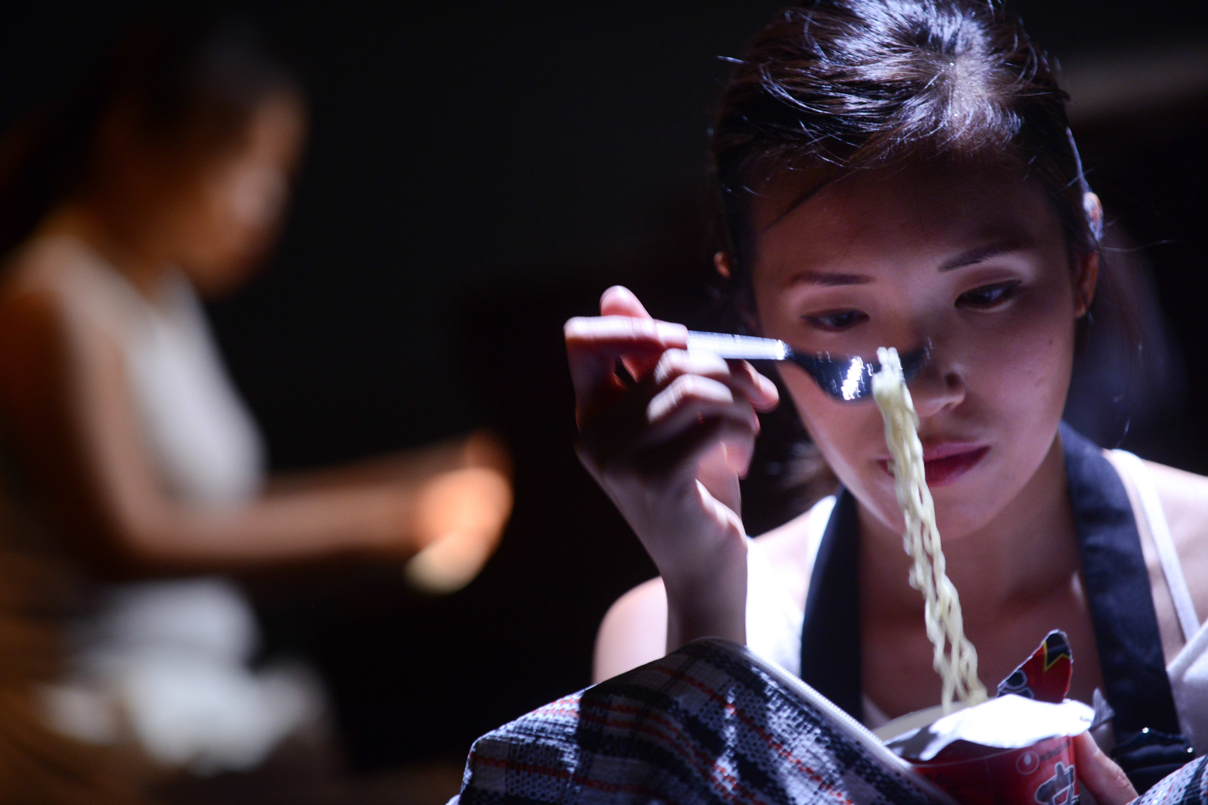 在這張來自天台製作（Rooftop Productions）當代香港戲劇《不是女僕》的劇照中，演員梁皓貽（Isabella Leung）置身於昏暗的舞台燈光下，展現出充滿張力的戲劇時刻。她神情專注地拿著一杯杯麵，用叉子挑起麵條，陰影下的光影勾勒出她沉思的神情，深刻呈現了演出中那份貼近日常的真實感。當角色回憶起這段相處時，她說道：「他知道我有一些杯麵，然後他會拿來吃。」