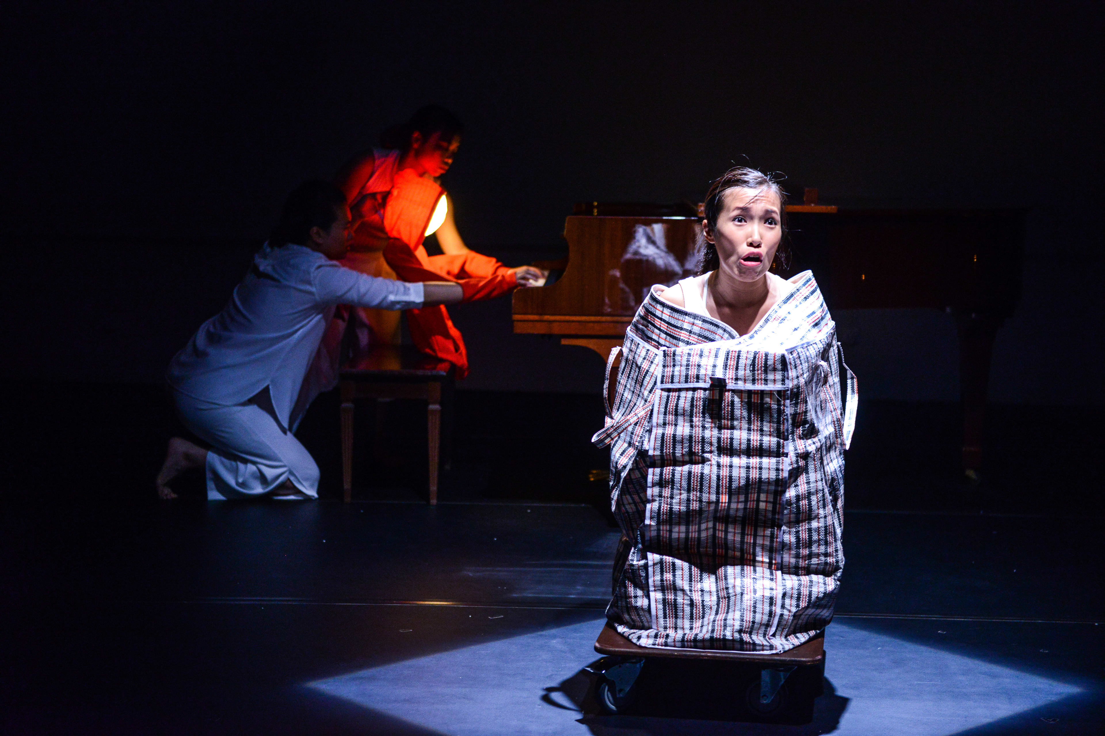在天台製作的當代劇場作品《不是女僕》中，演員梁皓貽（Isabella Leung）在戲劇性的一幕中呈現出極具張力的表演。她置身於一個巨大的紅白藍膠袋中，坐在一個帶輪的推車上，在聚光燈下流露出驚訝或困惑的神情。背景光線昏暗，另一位身著白色服裝的演員跪在鋼琴旁，而另一位身著橙色衣物的表演者則被一個發光的圓球照亮。這場香港實驗舞台劇深入探討了身份與家庭生活等議題。梁皓貽在劇中台詞道：「我們住在酒店，所以嚴禁養寵物。」