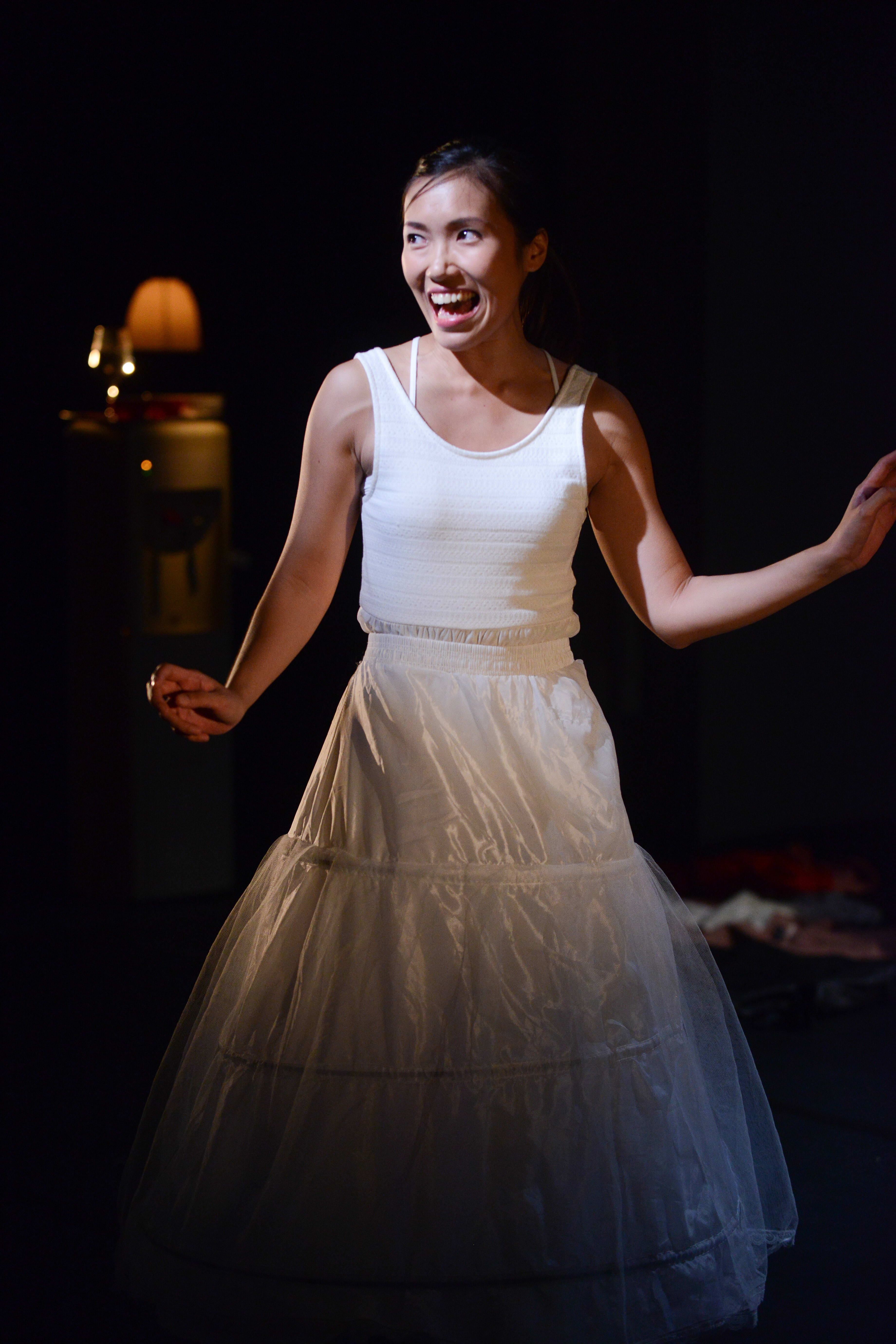 在天台製作 (Rooftop Productions) 當代香港劇場演出《不是女僕》(Not The Maids) 的戲劇性一幕中，演員梁皓貽站在舞台中央的聚光燈下。她身穿白色背心和蓬鬆的白紗裙，臉上帶著誇張、極具張力且帶點瘋狂的笑容。昏暗且極簡的舞台設計突顯了她的表演，捕捉到角色情緒激昂的瞬間。梁皓貽喊道：「還有你說的那間中介公司嗎？介紹我呀！」
