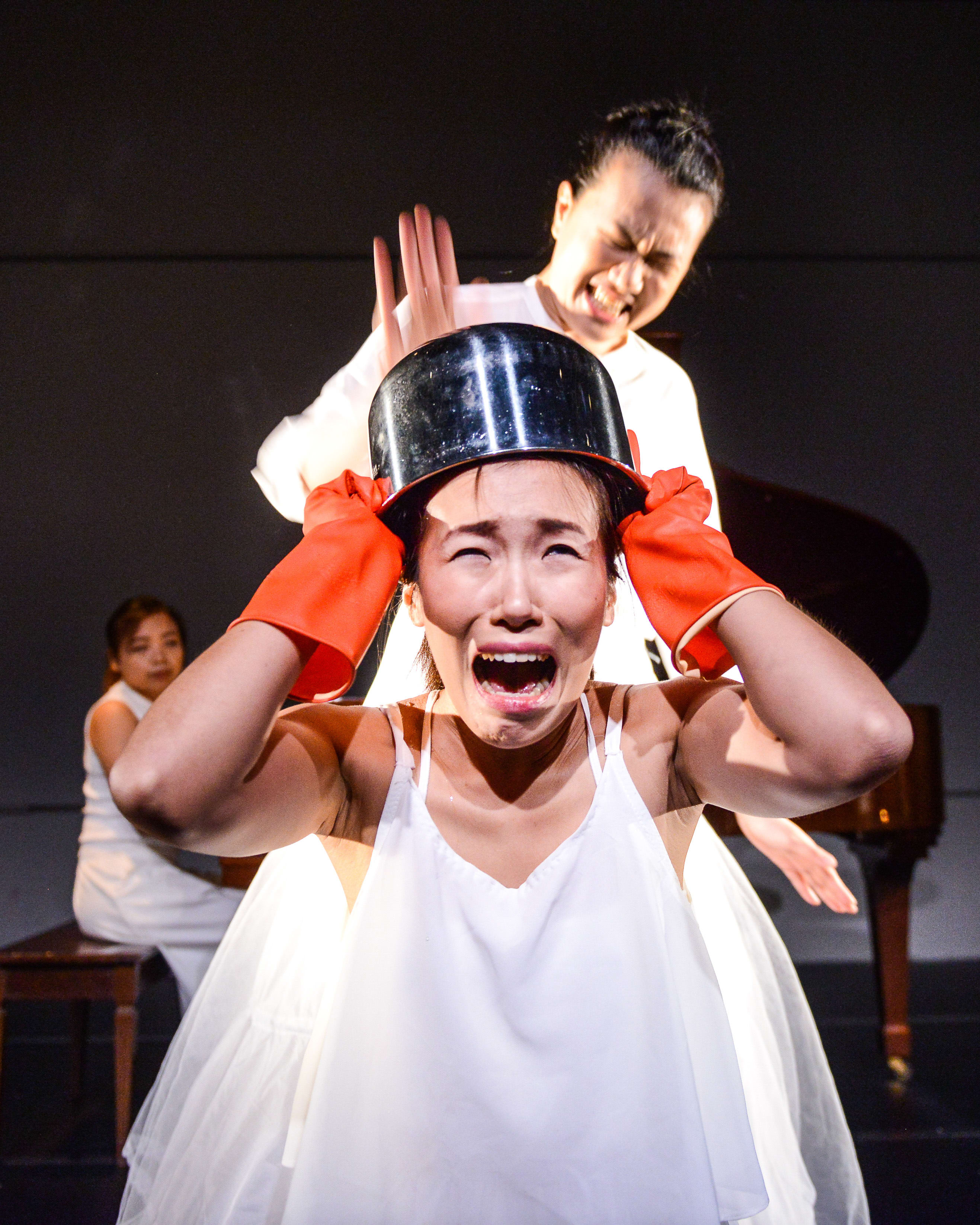 天台製作（Rooftop Productions）當代劇場演出《不是女僕》（Not The Maids）的戲劇張力時刻：演員梁皓貽頭戴金屬鍋，戴著紅色橡膠手套，表情痛苦且高聲呼喊；李婉晶則在其身後做出強烈的互動姿態。舞台背景昏暗，伴有鋼琴佈景，整體視覺呈現出關於家務勞動與身份認同的深刻隱喻。劇中角色悲憤地問道：「你說要太太洗，你，你覺得對不對？」