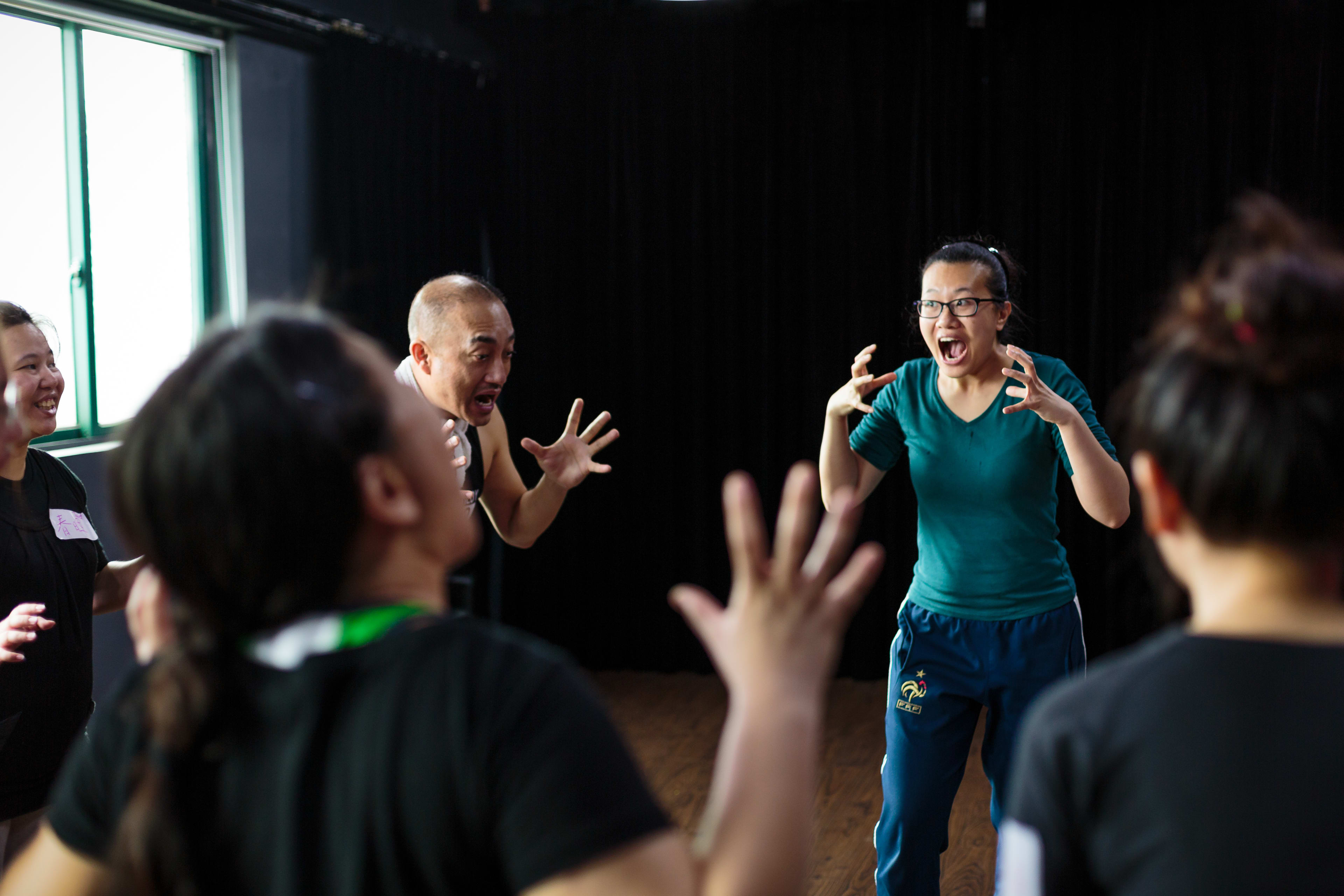 學員參與由天台製作（Rooftop Productions）舉辦的「廣州形體劇場及復調演唱工作坊」，正在進行情感豐富的肢體與聲音練習。導師李婉晶（Michelle Li）在中央示範充滿張力的發聲技巧，雙手舉起，神情投入，其他學員亦在排練室中熱烈呼應。廣州形體劇場及復調演唱工作坊。