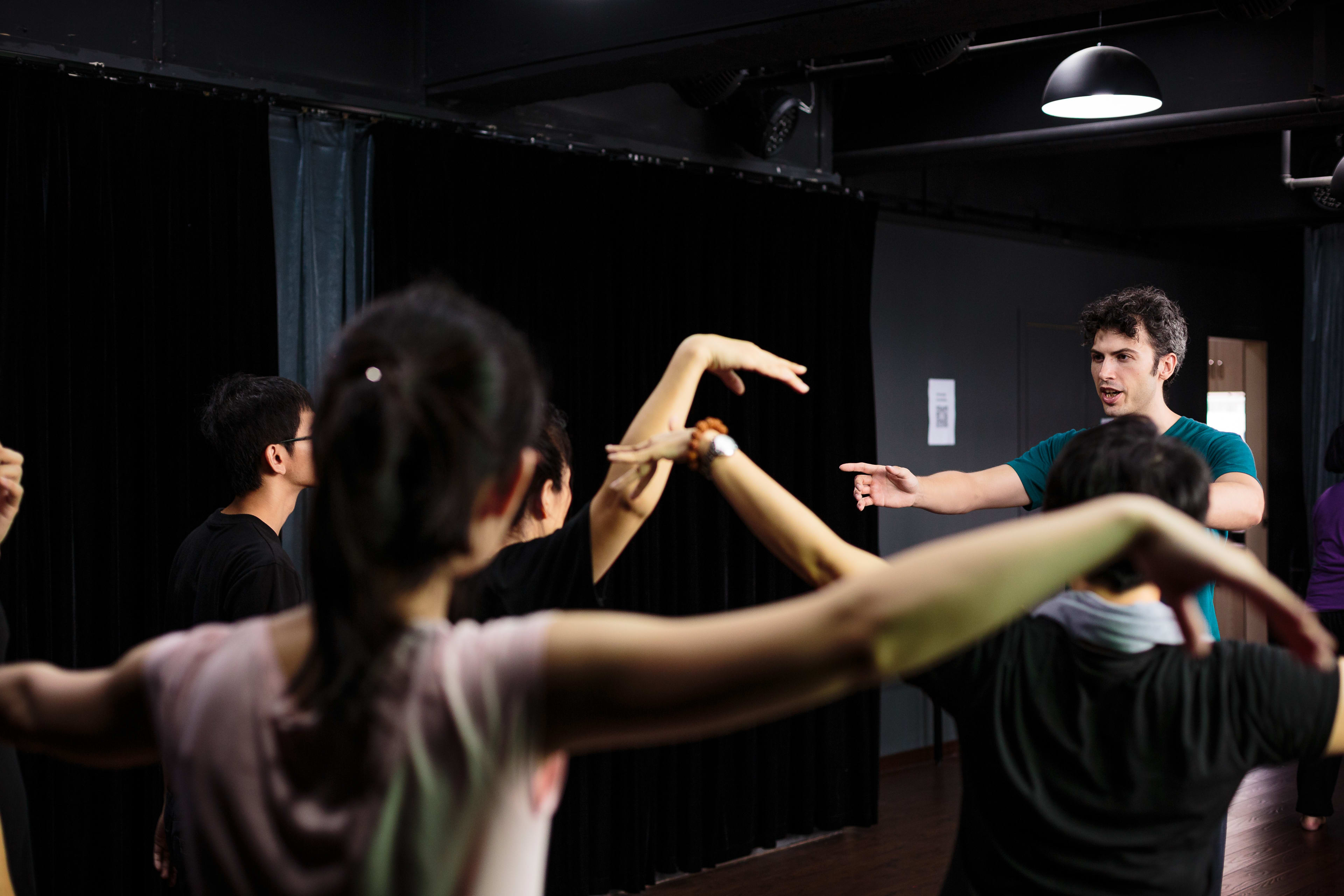 A candid shot taken during a Guangzhou Physical Theatre workshop hosted by Hong Kong-based Rooftop Productions. In a rehearsal space with dark curtains and dramatic overhead lighting, lead instructor Ivor Houlker (艾浩家) stands to the right, directing a group of performers who are practicing expressive arm movements and physical theatre techniques. The focus is on the dynamic, polyphonic nature of the movement, capturing the artistic process behind the Guangzhou Physical Theatre Final Performances.