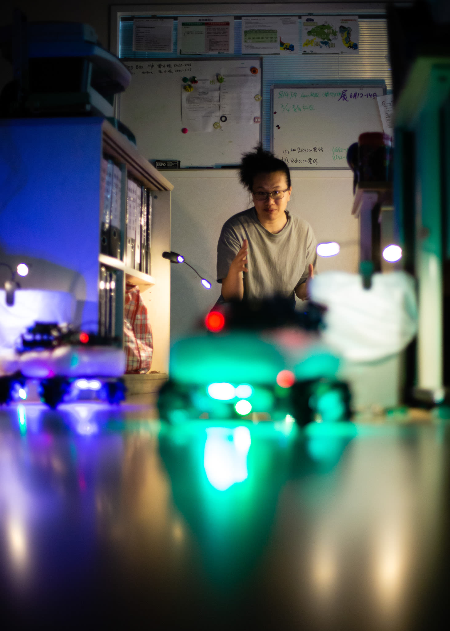 在天台製作（Rooftop Productions）當代劇場作品《遙距在場》（Presence）的排練中，演員李婉晶（Michelle Li）置身於昏暗的辦公室場景內。前景中一個小型機器人散發出模糊的綠色光芒，映照出充滿劇場感的氛圍。李婉晶眼神專注，手勢細膩，展現出與科技互動的戲劇張力。畫面呈現了她說道：「第二個任務：找到鑰匙。」