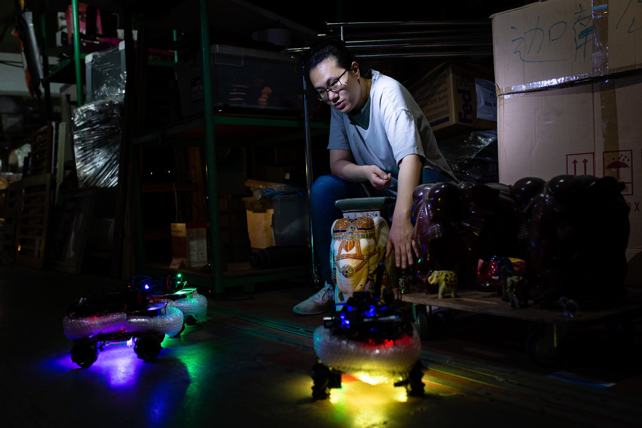 In a dim, atmospheric rehearsal space, performer Michelle Li sits on a wooden crate next to a decorative elephant sculpture, interacting with small, glowing remote-controlled toy vehicles on the floor. This rehearsal photo captures the experimental stagecraft of Rooftop Productions' (天台製作) contemporary theatre piece, 'Presence' (遙距在場), where mechanical elements meet human connection. As she gestures toward the illuminated toys, Michelle Li narrates, 'The elephant story'.