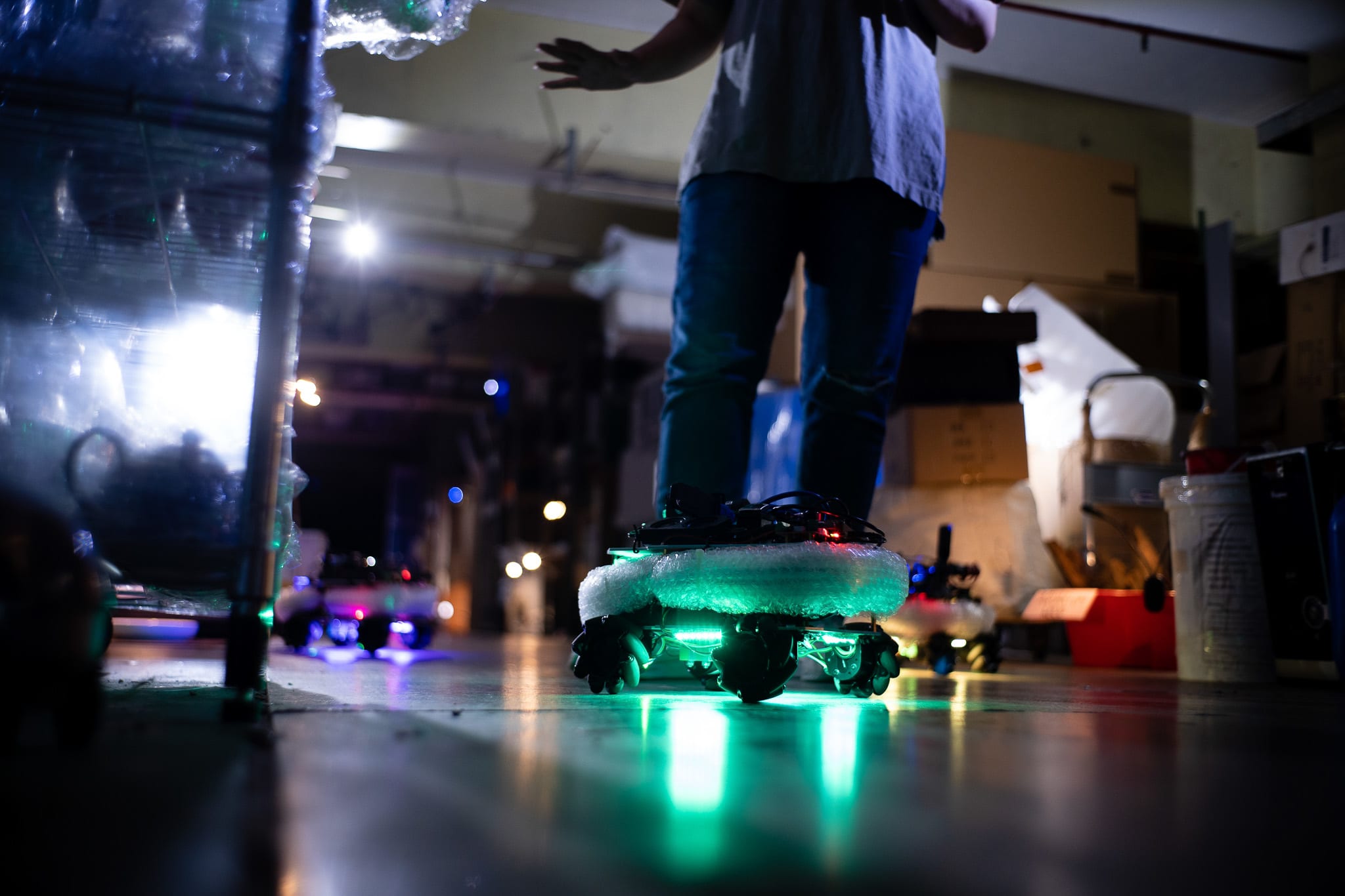 在這張天台製作（Rooftop Productions）當代劇場作品《遙距在場》（Presence）的排練照中，一位表演者站在一個散發著螢光綠LED燈光的四輪機器人平台上，場景位於昏暗的儲存區域。這畫面呈現了當代香港劇場的實驗性特質，結合了科技元素與親密的場域特定敘事。表演者正在探索儲存區域。「探索儲存區域」