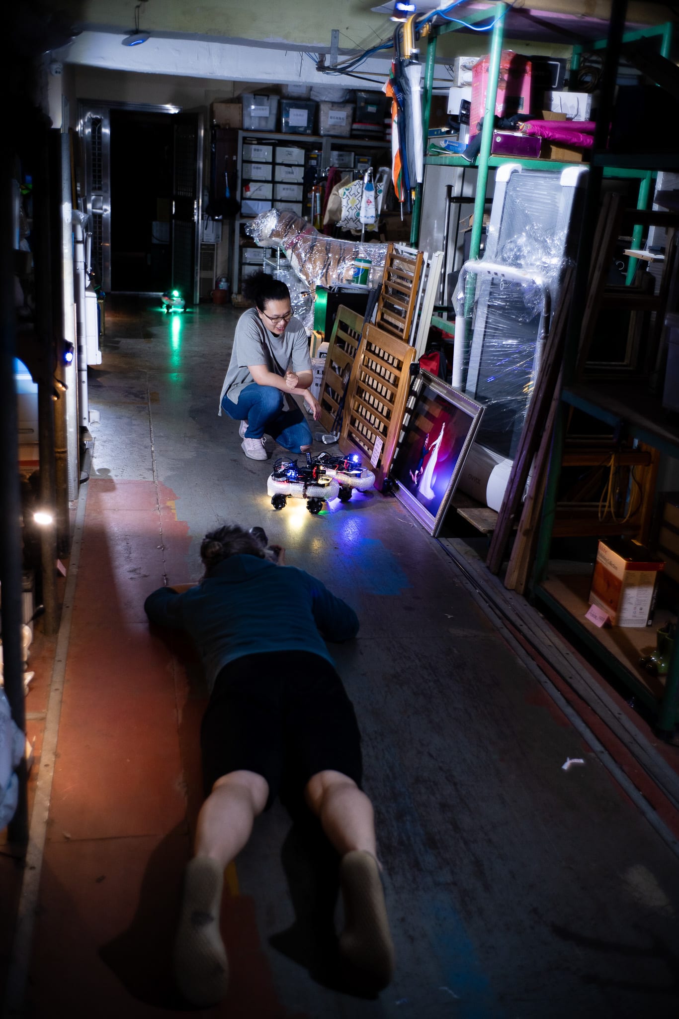 天台製作（Rooftop Productions）當代劇場作品《遙距在場》（Presence）的幕後排練花絮。在昏暗的儲物空間內，一位表演者趴在地上，為微型機械裝置拍攝低角度影像，李婉晶（Michelle Li）則在旁蹲守，神情專注地觀察着這場充滿實驗色彩的香港舞台製作過程。「拍攝中……」