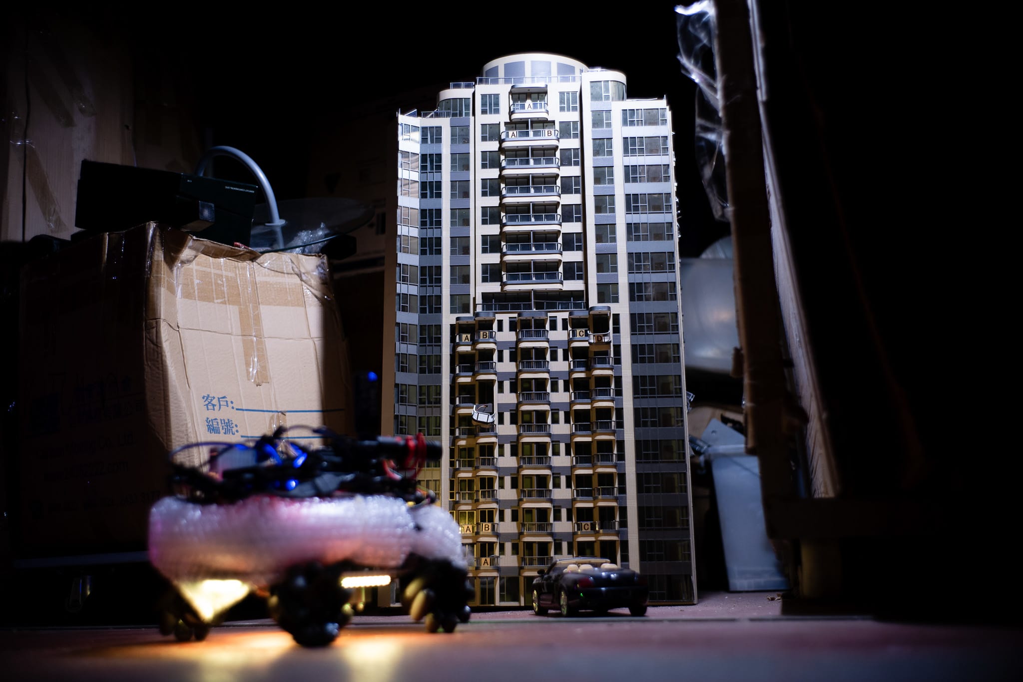 天台製作（Rooftop Productions）當代劇場演出《遙距在場》（Presence）的舞台佈景。畫面中是一座精緻的香港高層住宅模型，在昏暗的空間裡被冷調燈光照亮。前景中，一個被氣泡紙包裹的遙控機械裝置與模型底部的玩具汽車形成呼應，隱喻城市空間中的疏離與遙距感。「香港的公屋」