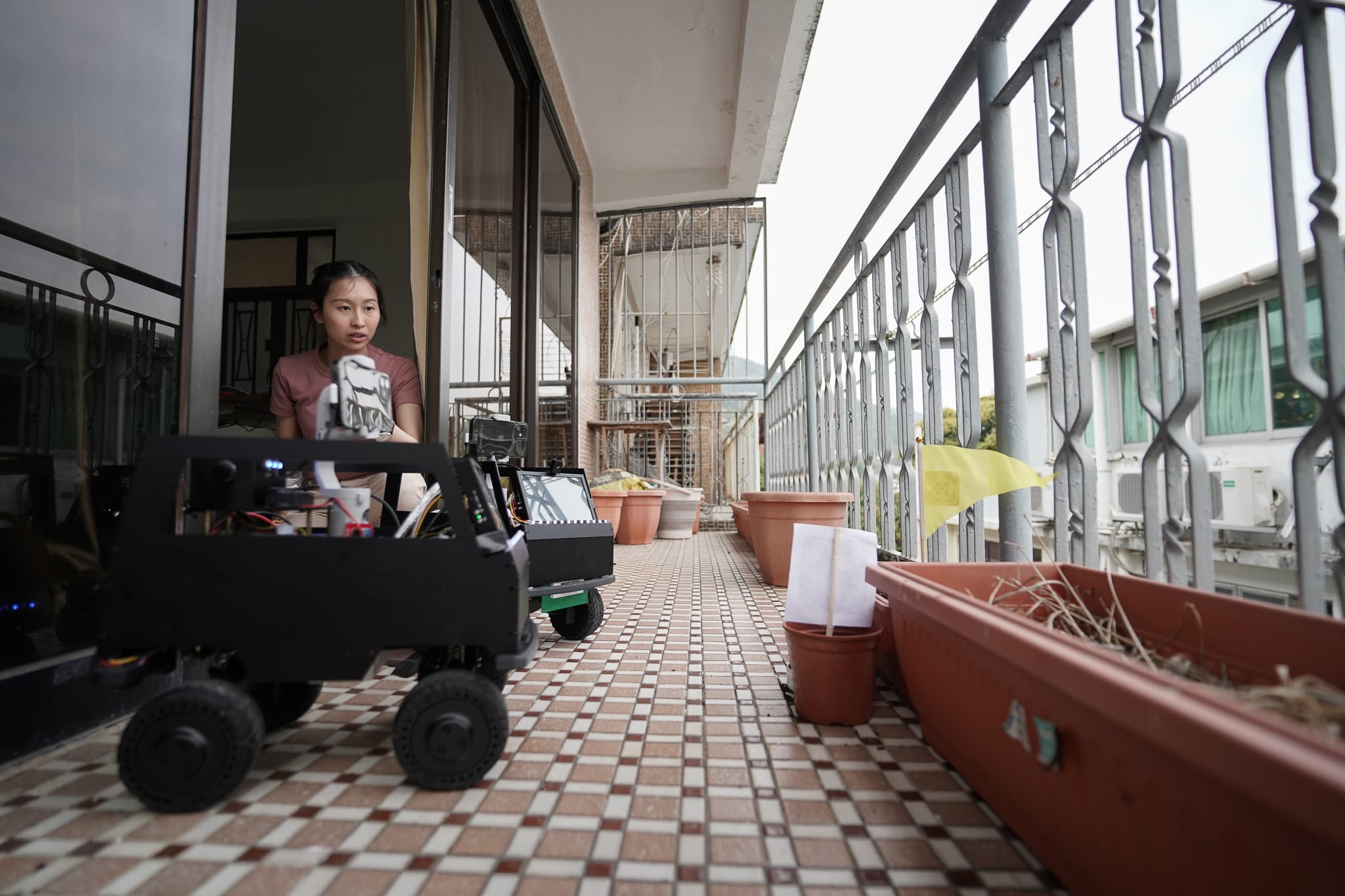 天台製作（Rooftop Productions）當代劇場作品《遙距在場》（Presence）劇照。演員蘇巧珊在住宅露台操作一部裝有鏡頭的黑色小型機械車，將科技元素融入私密的敘事表演中。在自然光影下，她神情專注地說道：「28歲生日正日我去咗睇醫生，因為全身都出咗啲紅斑。」