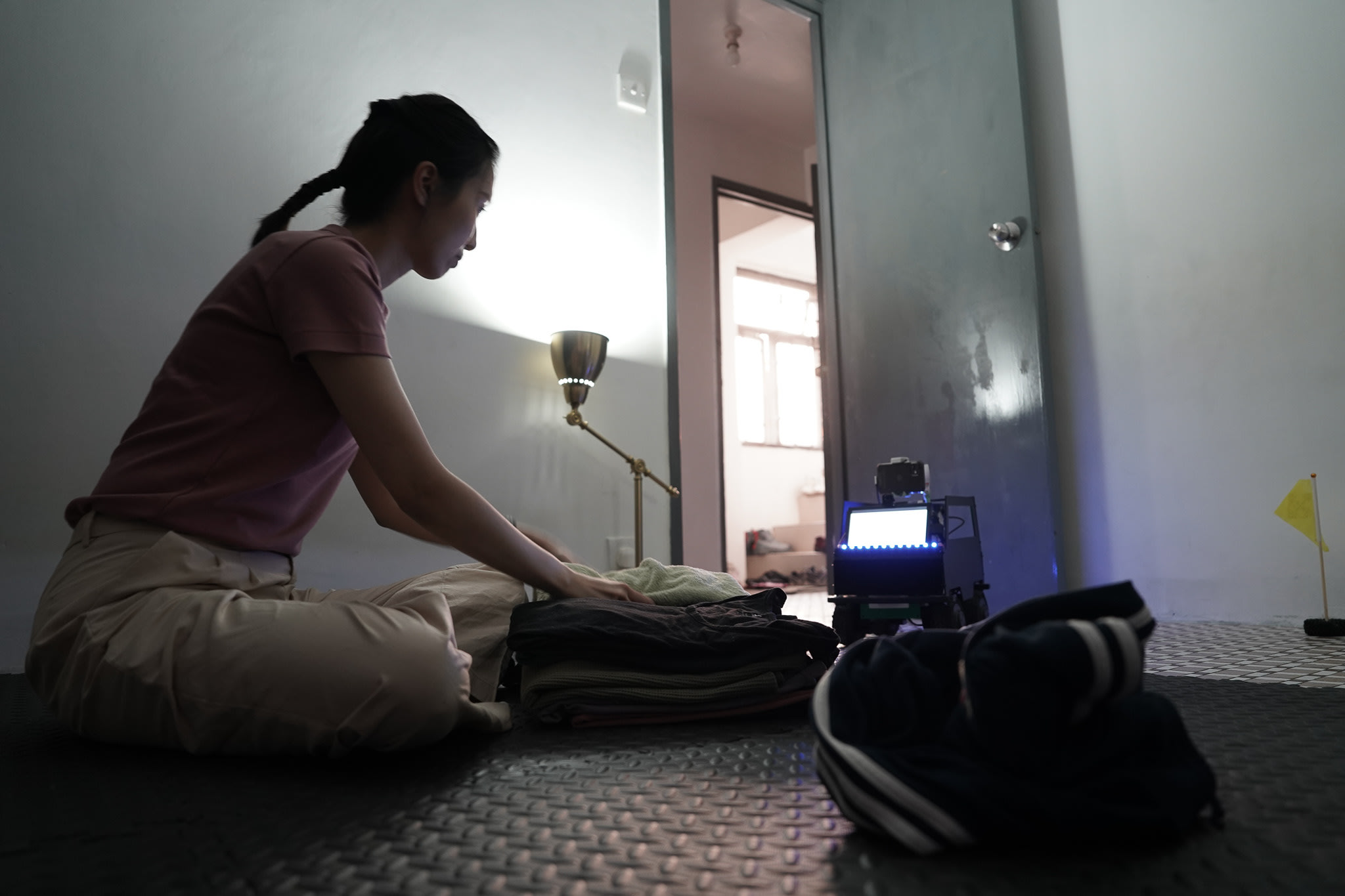 在天台製作的當代劇場作品《遙距在場》（Presence）排練中，演員蘇巧珊盤腿坐在鋪設膠墊的暗室內。她一邊專注地摺疊衣物，一邊與身旁發著微光的機械裝置進行互動。這幕場景呈現了該劇在科技與人性之間獨特的思考，正如她所言：「仲甩緊.......好多頭髮........剩番一成都冇。」
