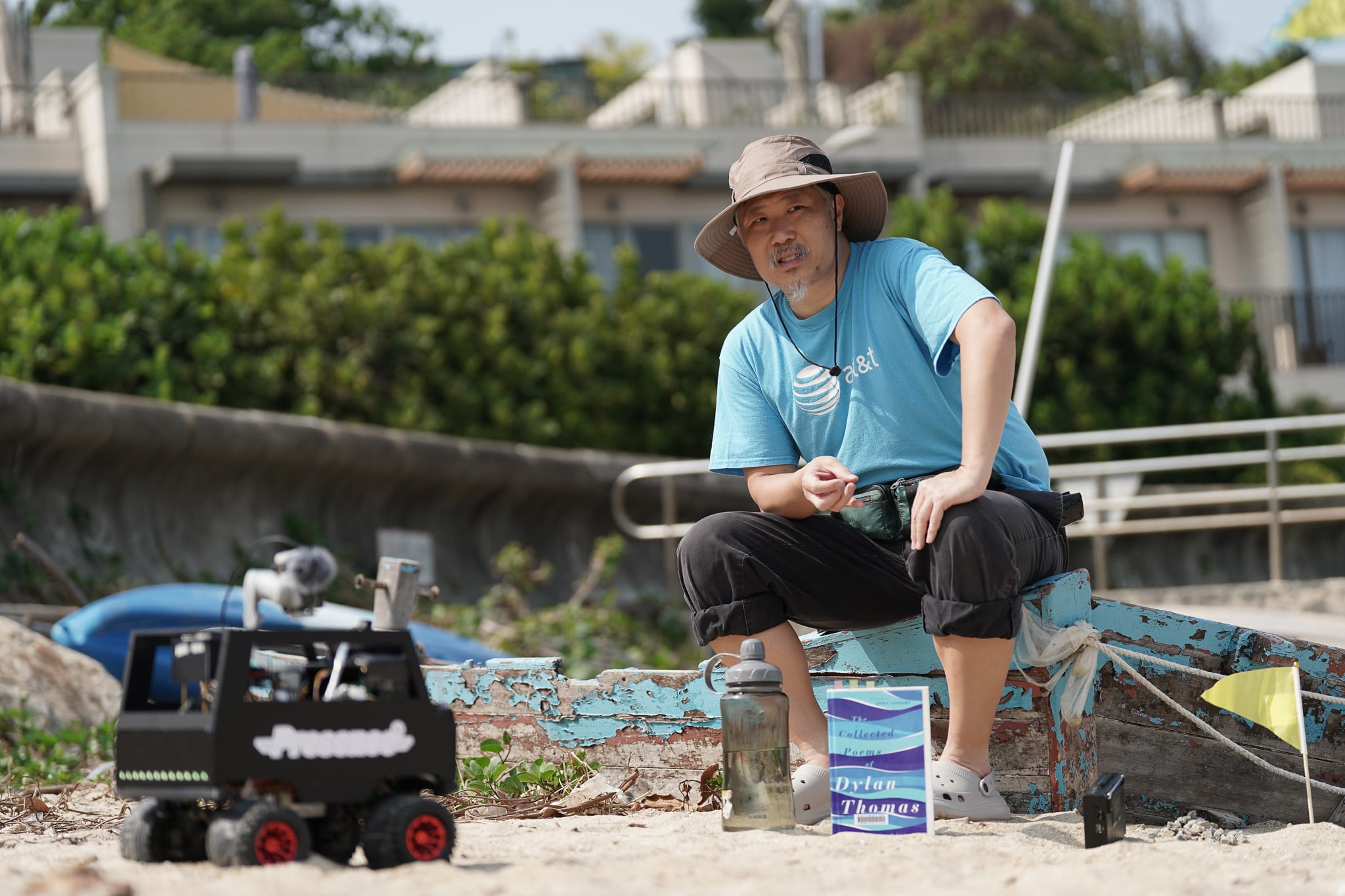宋本浩坐在沙灘上一艘斑駁的破舊木船上，參與天台製作（Rooftop Productions）當代劇場作品《遙距在場》（Presence）的排練。他頭戴遮陽帽，身穿藍色T恤，正凝視著沙灘上的一台黑色小型遙控機器人。他身旁擺放著水瓶和一本《戴蘭·湯瑪斯詩集》，為這部實驗性舞台演出增添了文學氣息。他若有所思地問道：「100年後會係點？」