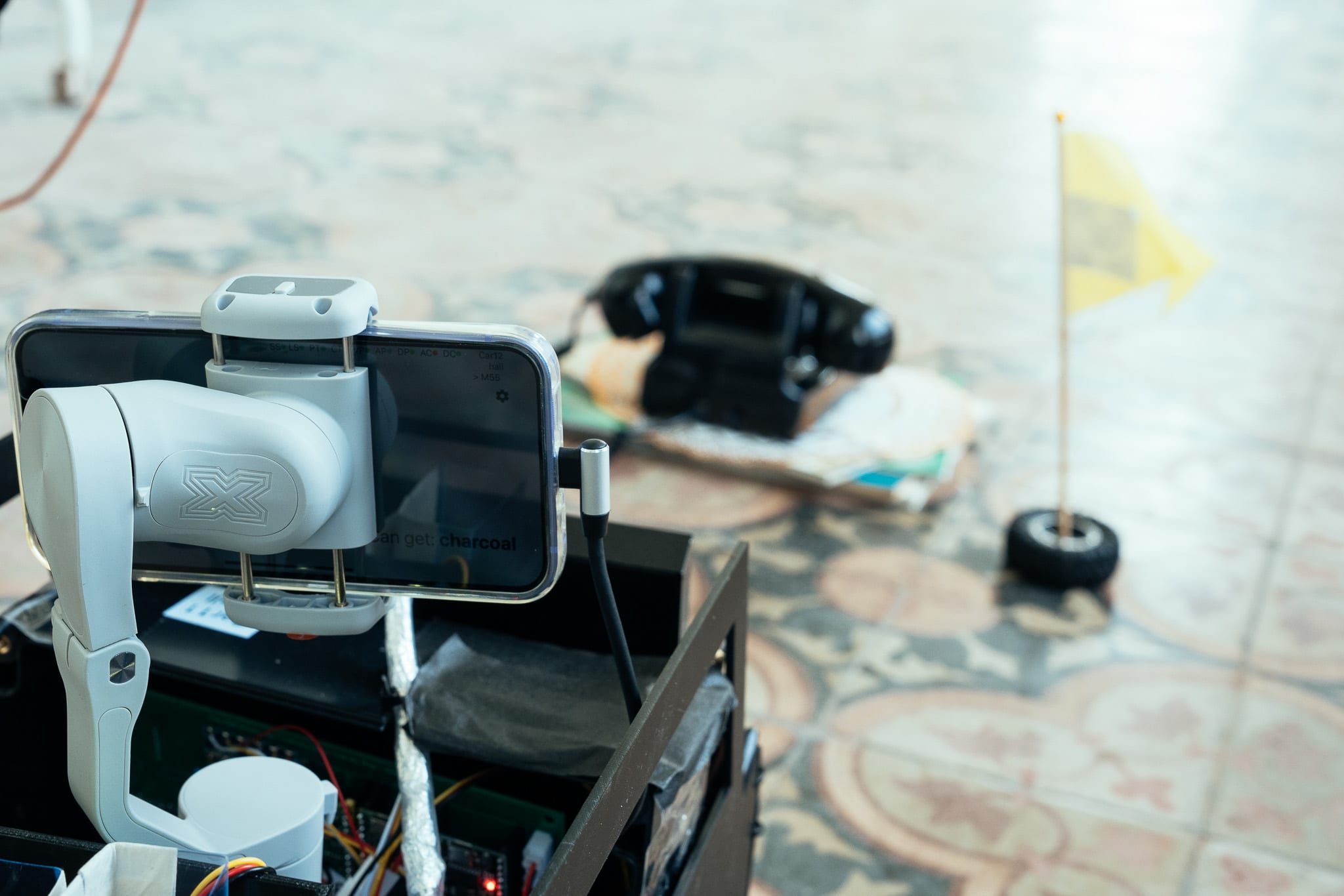 A close-up view of a smartphone mounted on a robotic gimbal, capturing a scene for Rooftop Productions' contemporary Hong Kong theatre performance 'Presence'. The camera points toward a vintage black telephone sitting on the floor in the background next to a small, yellow marker flag, evoking the nostalgic atmosphere of the production. 'There was a period of time when Sam So was the receptionist in U hall and she was responsible for the only telephone there.'