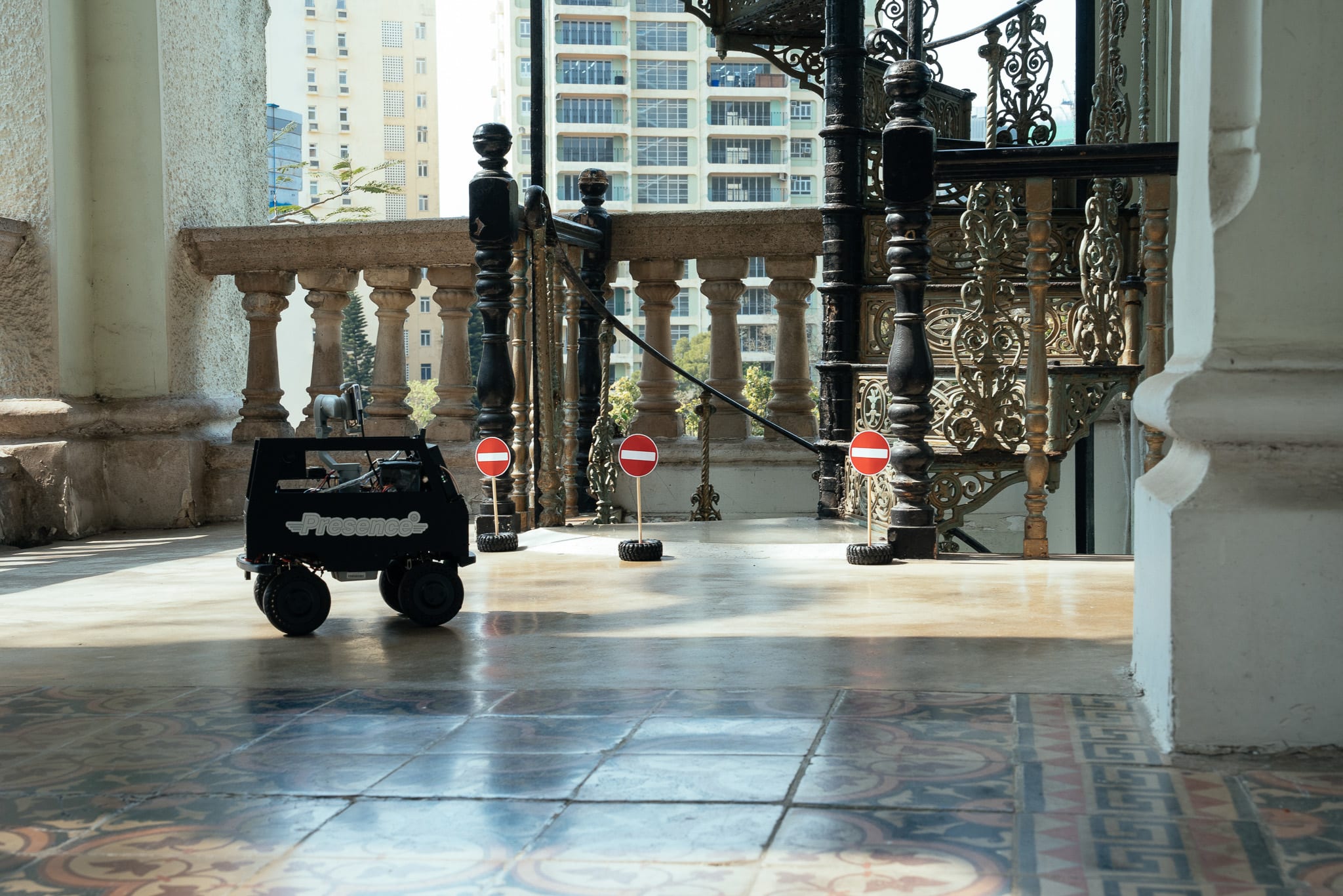 A small, black, toy-like vehicle labelled 'Presence' sits on a polished, patterned tiled floor of a historic building in Hong Kong, positioned before three miniature 'no entry' road signs. The scene, evocative of Rooftop Productions' (天台製作) experimental theatre work, captures a moment from the production 'Presence' (《遙距在場》), where architectural history meets contemporary performance. 1954 - the University purchased the property from the French Mission at the price of 1.6 million Hong Kong Dollars.