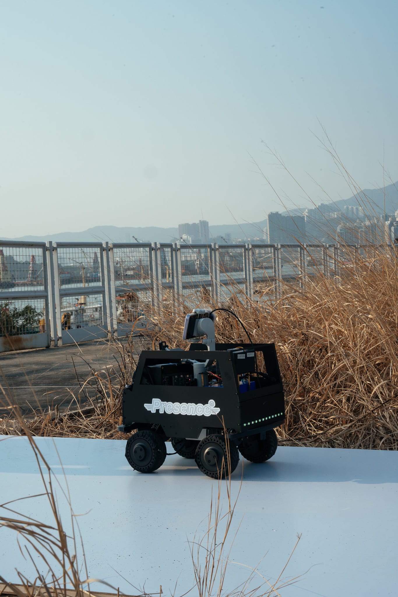 一台印有「Presence」字樣的黑色遙控機械車停在白色地面與草叢之間，車身安裝的攝影機正在進行拍攝。背景為鐵絲網與香港工業區的天際線，呼應天台製作（Rooftop Productions）當代劇場演出《遙距在場》的視覺風格。天際觀景台。