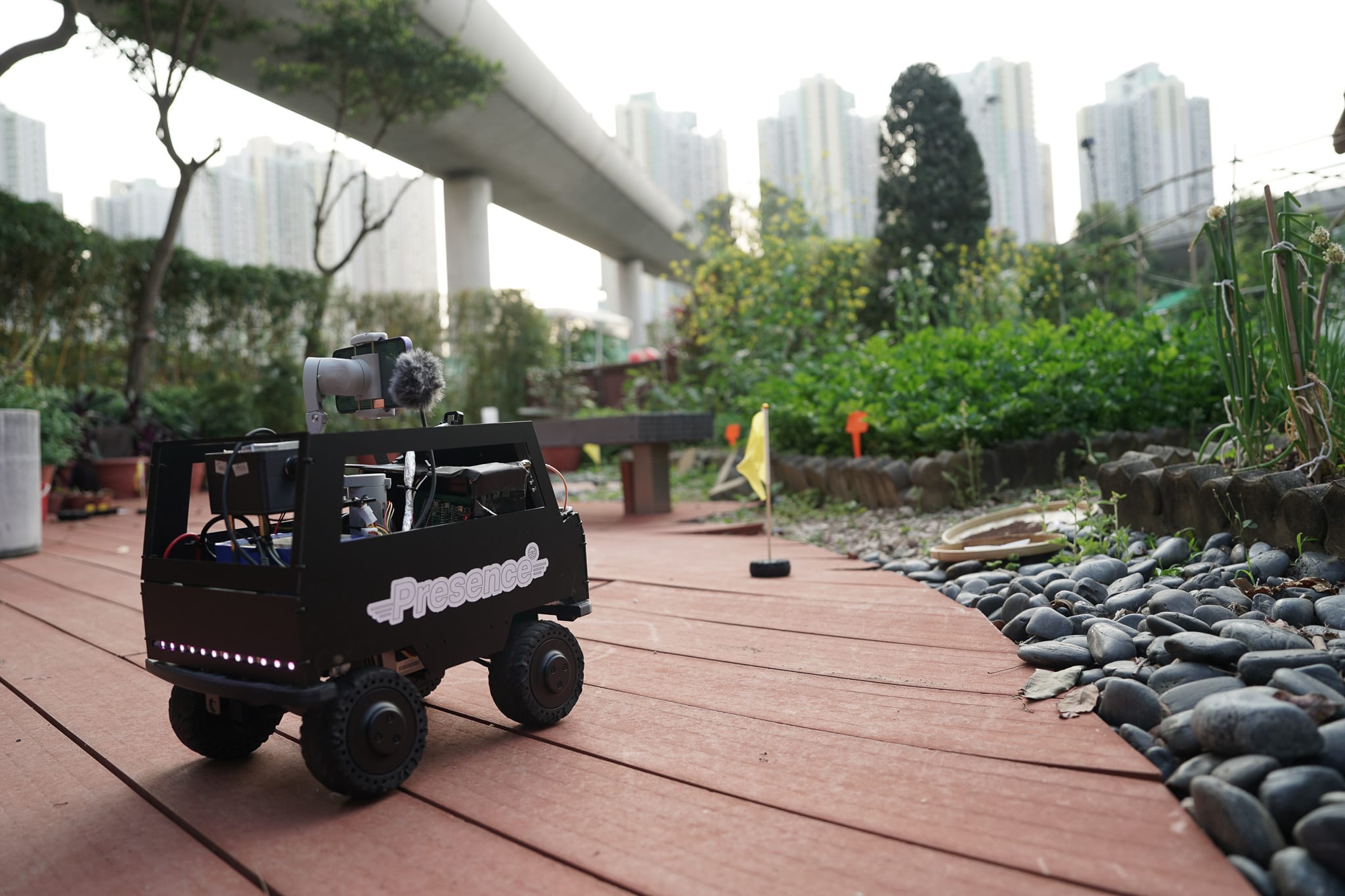 一台印有 'Presence' 字樣的黑色遙控機器車，正行駛在香港天台花園的木棧道上。該車配備雲台攝像機，呈現天台製作（Rooftop Productions）當代劇場作品《遙距在場》（Presence）的排練或演出現場。背景映襯著現代住宅大廈，突顯了科技與人類居住環境之間的微妙張力。「人類需要多少土地？」