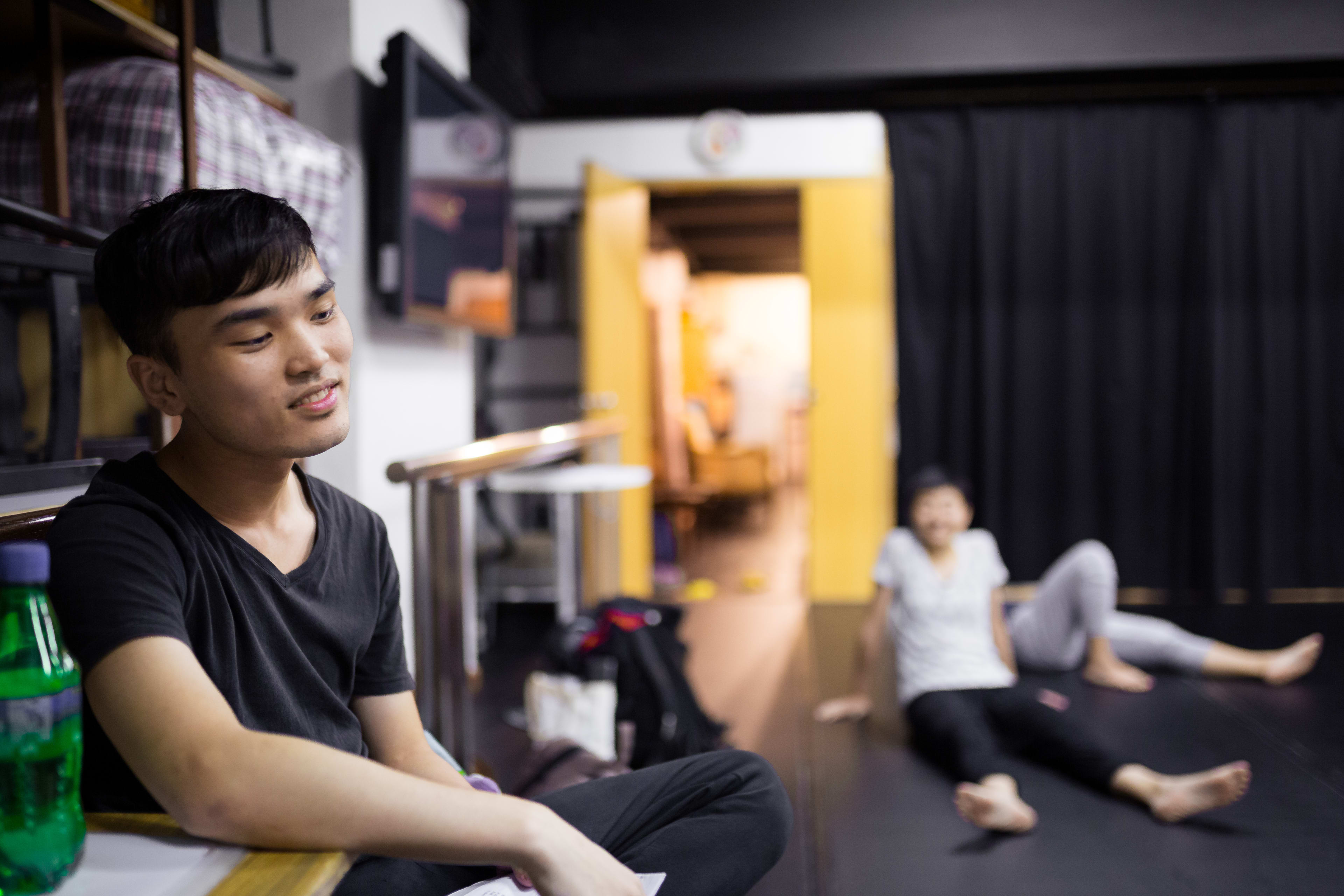 在一間光線明亮的排練室中，一位年輕男演員坐在前景，在參與當代劇場工作坊的休息期間若有所思地看向旁邊。背景中，另一位學員模糊地坐在地板上休息，展現出這場於香港舉辦的「身心合一」(Psychophysical Actor Training) 工作坊中，既放鬆又專注的排練氛圍。Psychophysical actor training workshops in Hong Kong, 2016.