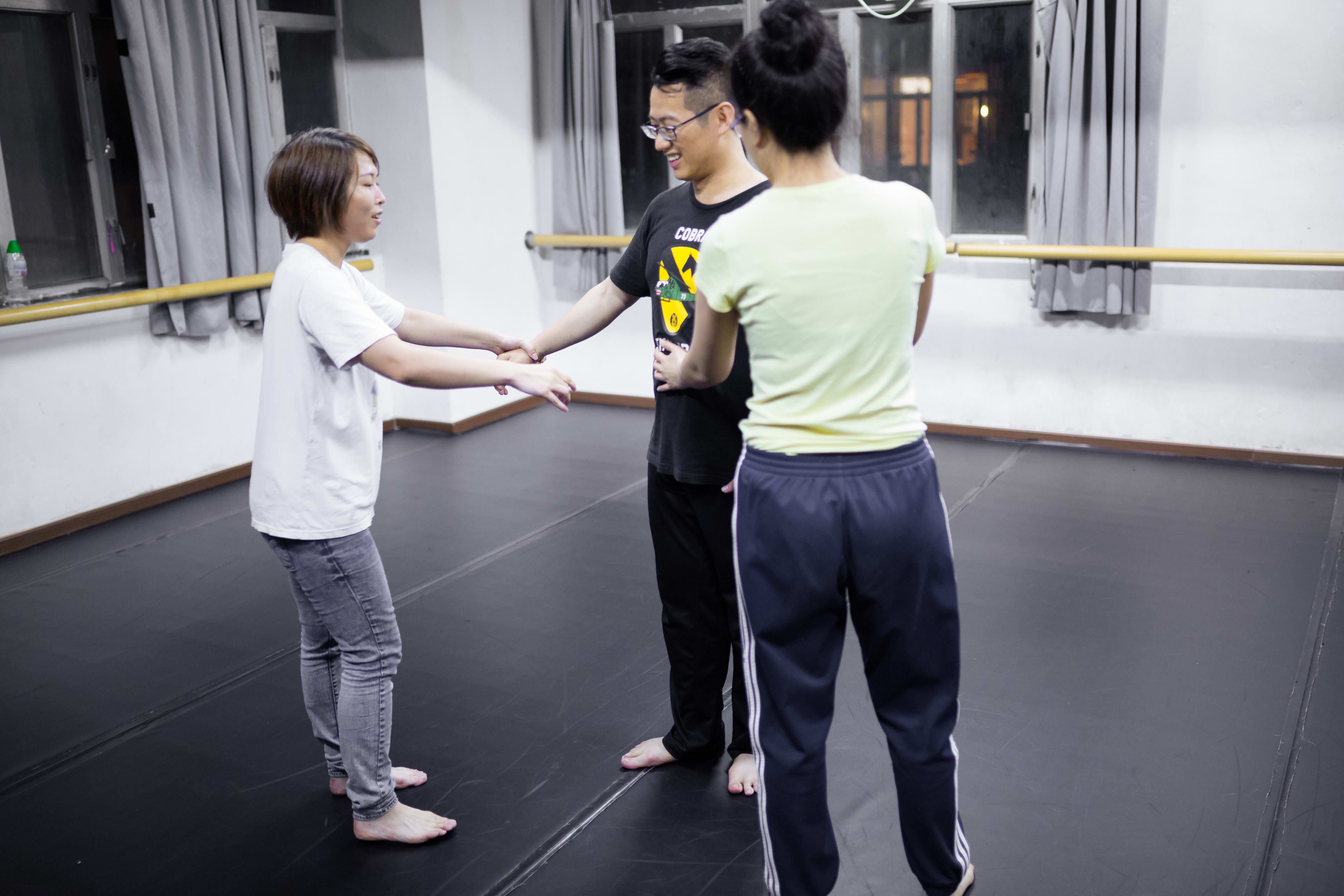 三位演員正在香港參與「身心合一」表演訓練工作坊。照片中，左右兩側的女性學員正引導中間的男性學員進行肢體與呼吸的練習，展現出天台製作（Rooftop Productions）對於當代劇場表演訓練的專注與細膩。場景設於設有芭蕾扶手與黑色地板的排練室中。Psychophysical actor training workshops in Hong Kong, 2016.