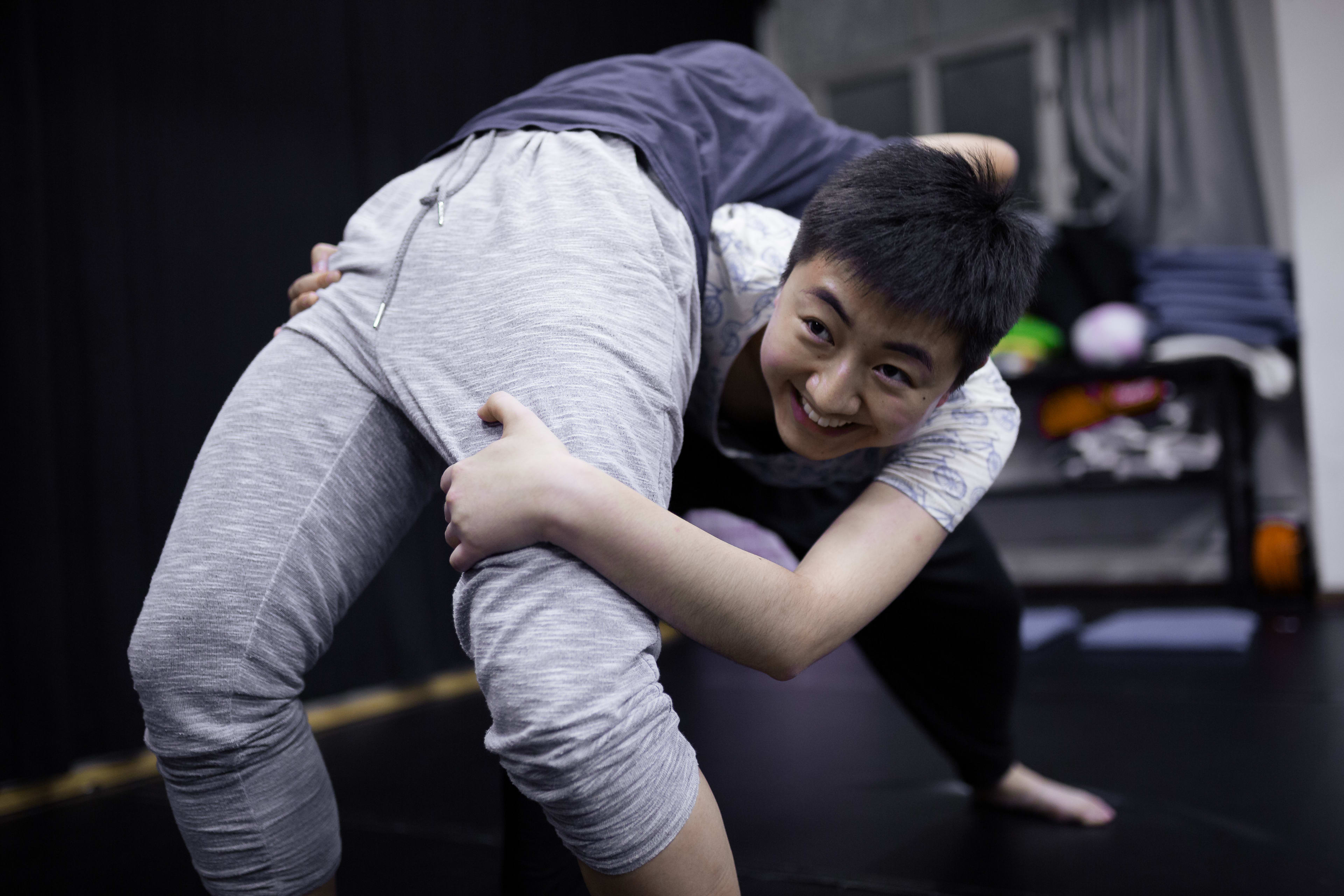 一名學員在香港「身心合一」表演工作坊中展示極致的身體靈活性，在練習過程中向後彎腰並伸手穿過雙腿，臉上帶著專注且開朗的笑容，體現了當代劇場對身心合一訓練的追求。Psychophysical actor training workshops in Hong Kong, 2016.