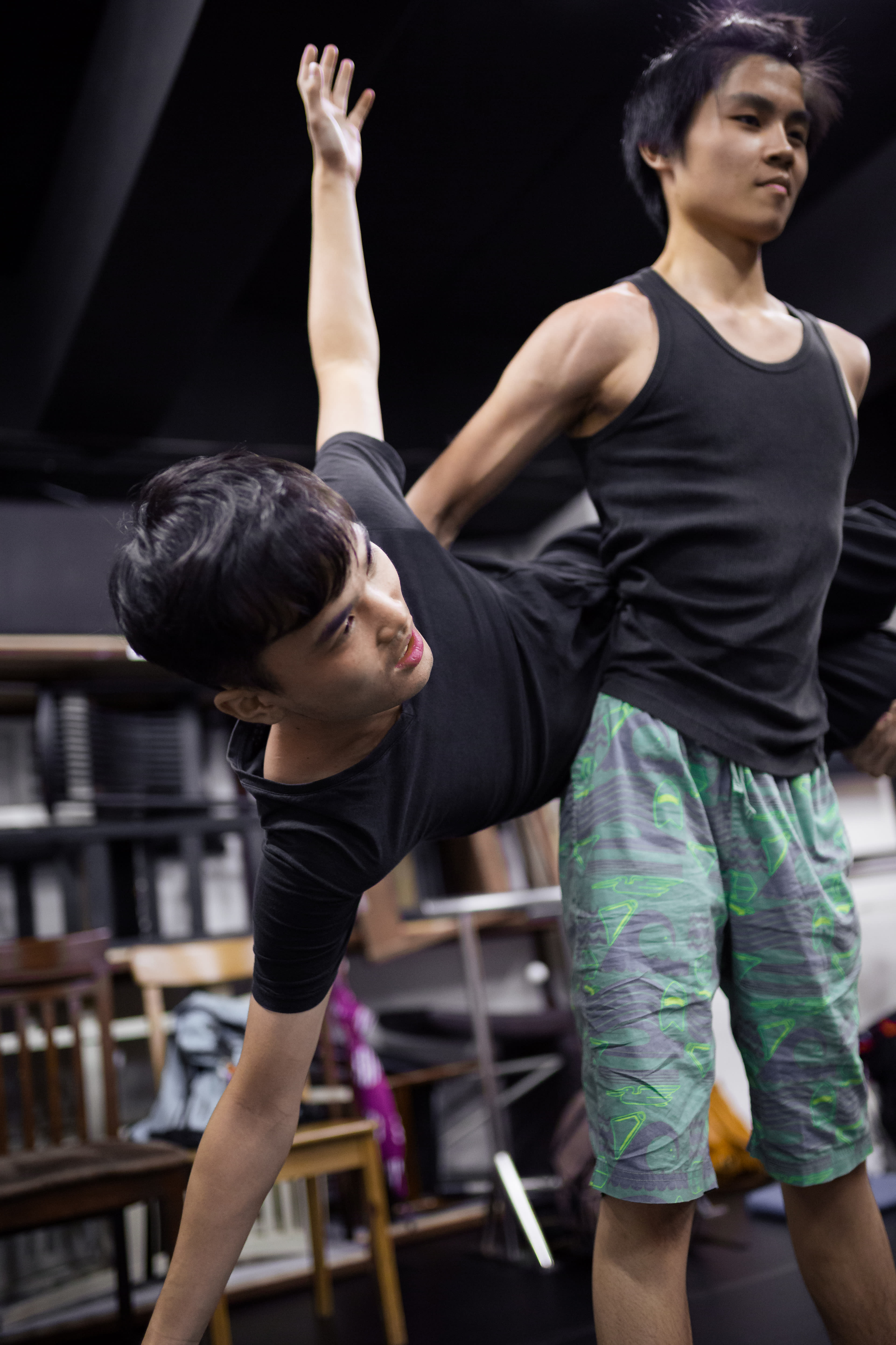 兩位演員正在香港參與「身心合一」（Psychophysical Actor Training）演員訓練工作坊。其中一位演員向側面大幅度傾斜，由另一位演員從旁協助，展現出天台製作（Rooftop Productions）表演訓練中極為講究的肢體掌控力與空間感。在排練室的燈光下，兩位演員展現出專注的神情，實踐肢體劇場的訓練技巧。Psychophysical actor training workshops in Hong Kong, 2016.