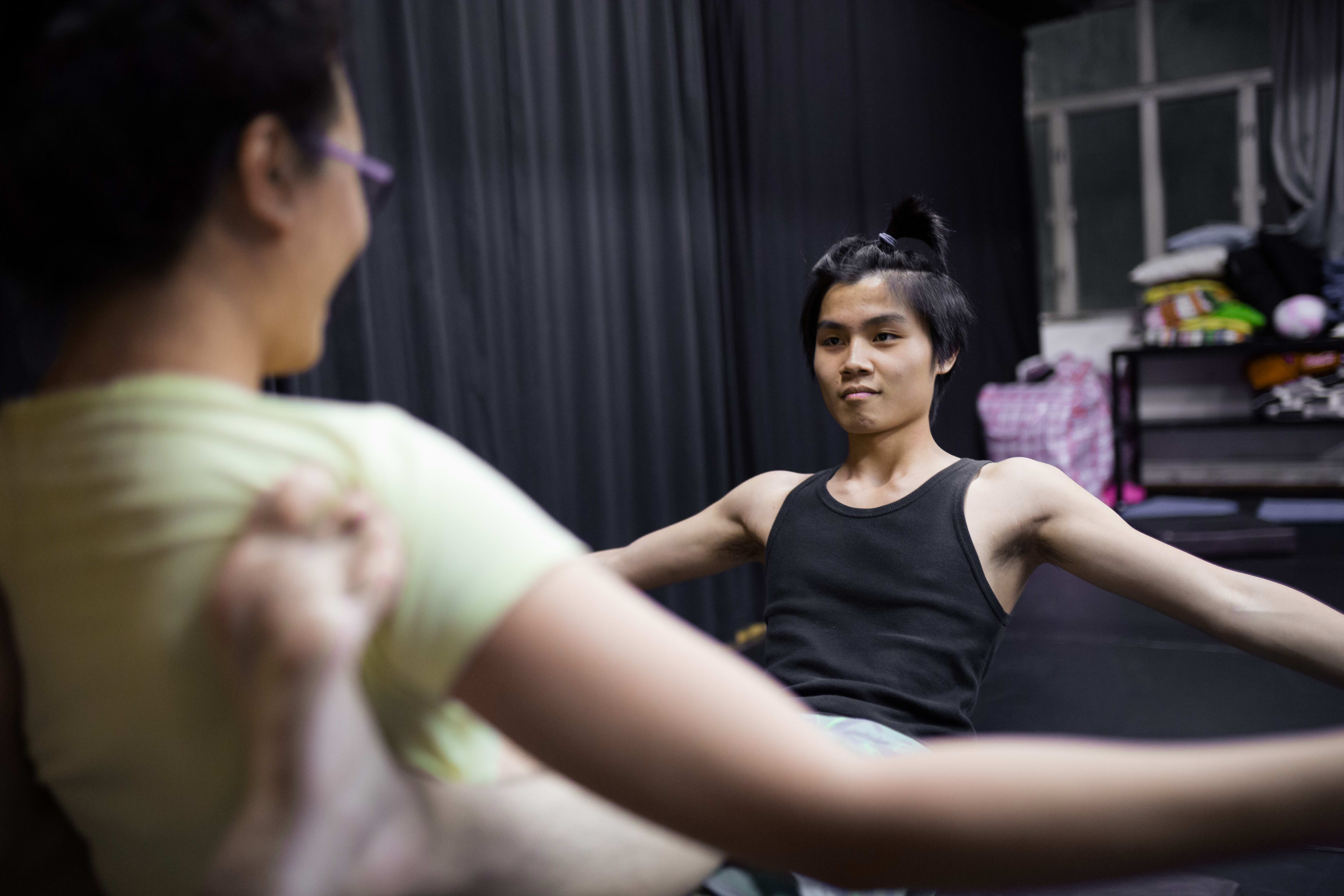 攝於 2016 年香港的「身心合一」演員訓練工作坊。畫面前景為伸展中的肢體，虛化處理帶出空間感；後方一名穿著黑色背心的年輕男演員正專注地進行核心訓練。場地以深色布幕為背景，呈現出香港當代劇場排練室的專業氛圍。「Psychophysical actor training workshops in Hong Kong, 2016」