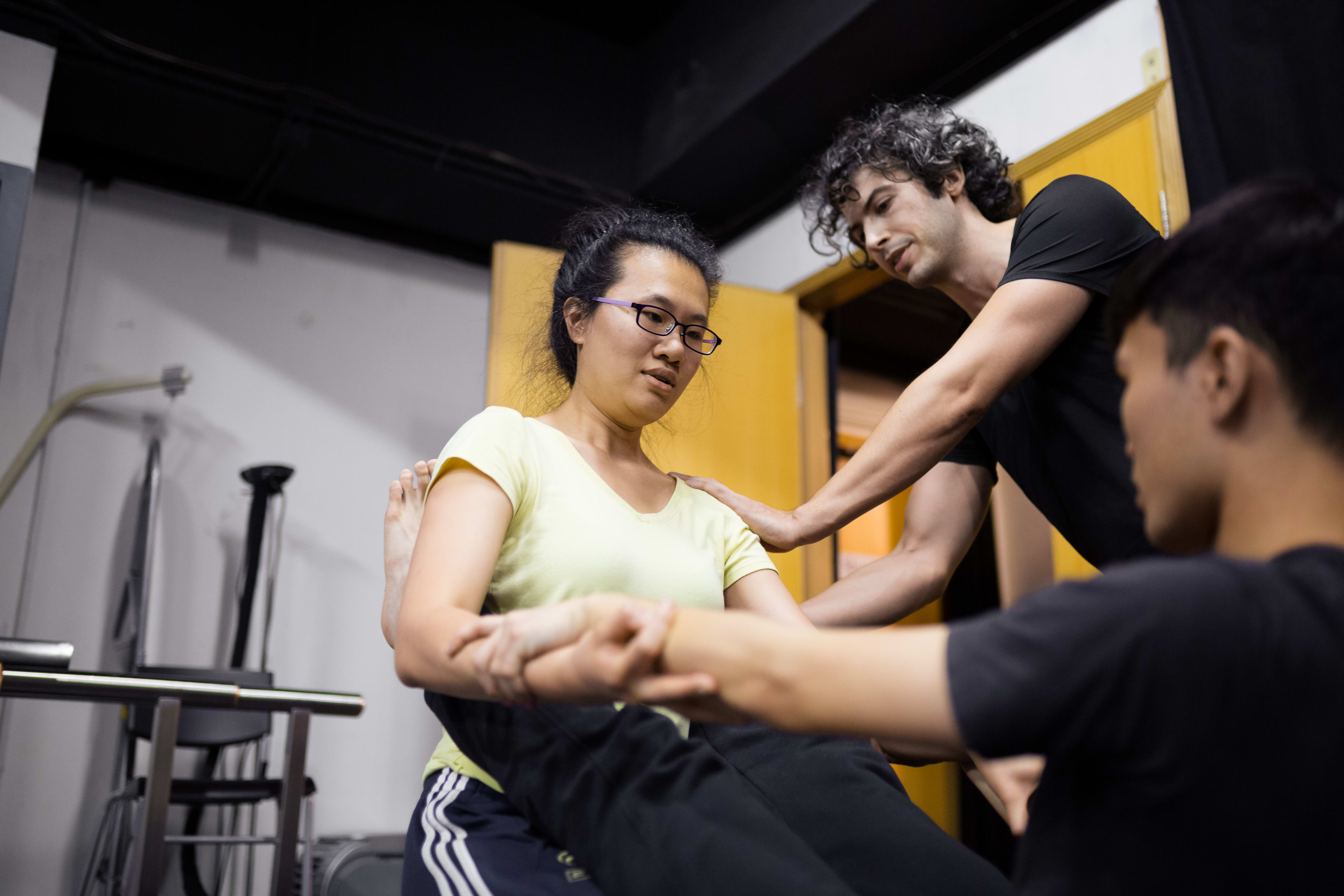在這張天台製作（Rooftop Productions）「身心合一」（Psychophysical Actor Training）工作坊的排練照中，演員李婉晶（Michelle Li）正坐在練習墊上，兩位導師正透過身體引導協助她探索空間感知與肢體控制。此照片攝於 2016 年的香港，展現了當代劇場訓練中嚴謹而專注的過程，體現了「身心合一」的表演理論。"Psychophysical actor training workshops in Hong Kong, 2016"