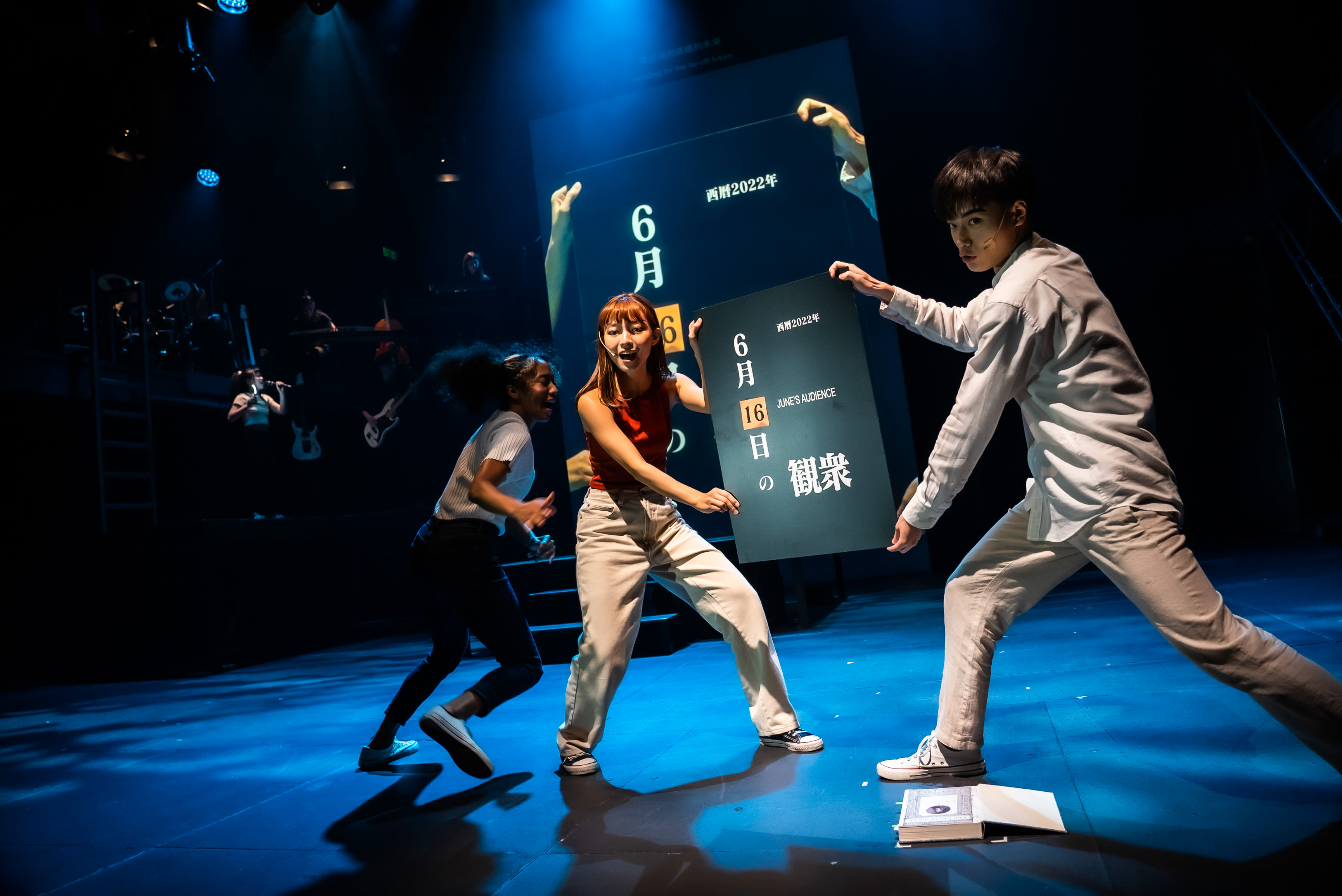 「天台製作」的當代香港劇場作品《天真與世故之歌》的一個充滿活力的場景。在戲劇性的藍色燈光下，三位表演者在昏暗的舞台上互動。中央，身穿紅色上衣的李安生（飾演摩洛）和淺色襯衫的羅文澤（飾演上帝）各自舉起黑板，上面顯示「6月16日 の観察」。左側，身穿白色上衣的冼樂欣（飾演夏娃或天使）正充滿能量地移動。後方大屏幕亦顯示著「西曆2022年」及類似文字，遠處的樂器設備暗示這是一場現場舞台演出。各位安坐於觀眾席的先生女士們。