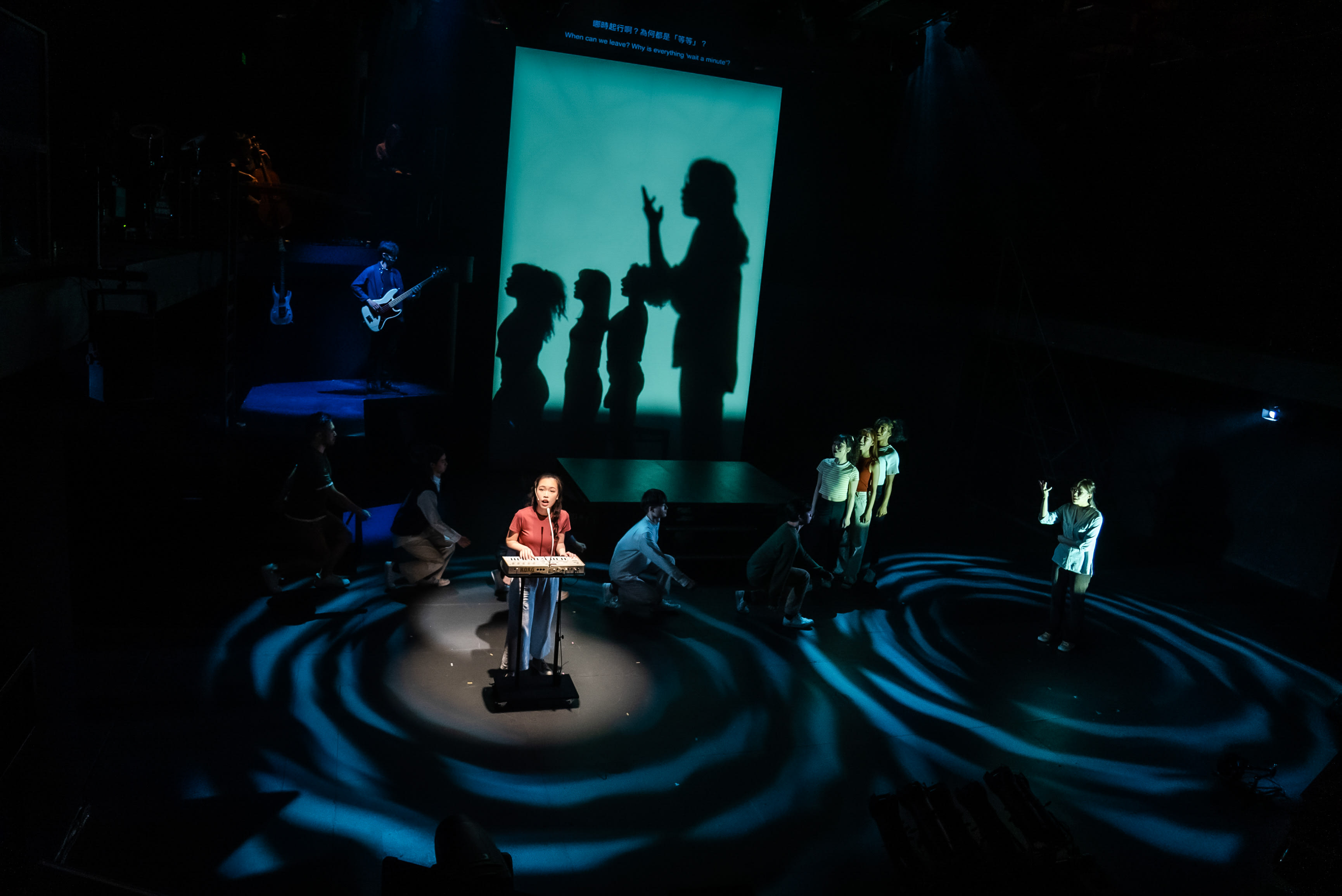 A dramatic contemporary Hong Kong theatre scene from Rooftop Productions' "Songs of Innocence and Experience" unfolds on a dark stage. In the foreground, actor Lee On-sang, in a red top and blue pants, stands at a black podium under a bright spotlight, her mouth open as if singing or speaking passionately. The stage floor is illuminated with swirling blue light patterns, creating a dynamic effect. To the right, actor Sin Lok-yan gestures upwards, while other cast members are dimly visible, standing and crouching. In the mid-ground on the left, Wu Lui-fung, the band leader, plays an electric guitar, bathed in blue light. A large, bright teal screen dominates the background, displaying dark silhouettes of several figures, including one with an arm raised, mirroring the pose of Sin Lok-yan, adding to the visual storytelling of this live stage performance. "Shum Cheuk Yiu, you don’t pick up my calls."