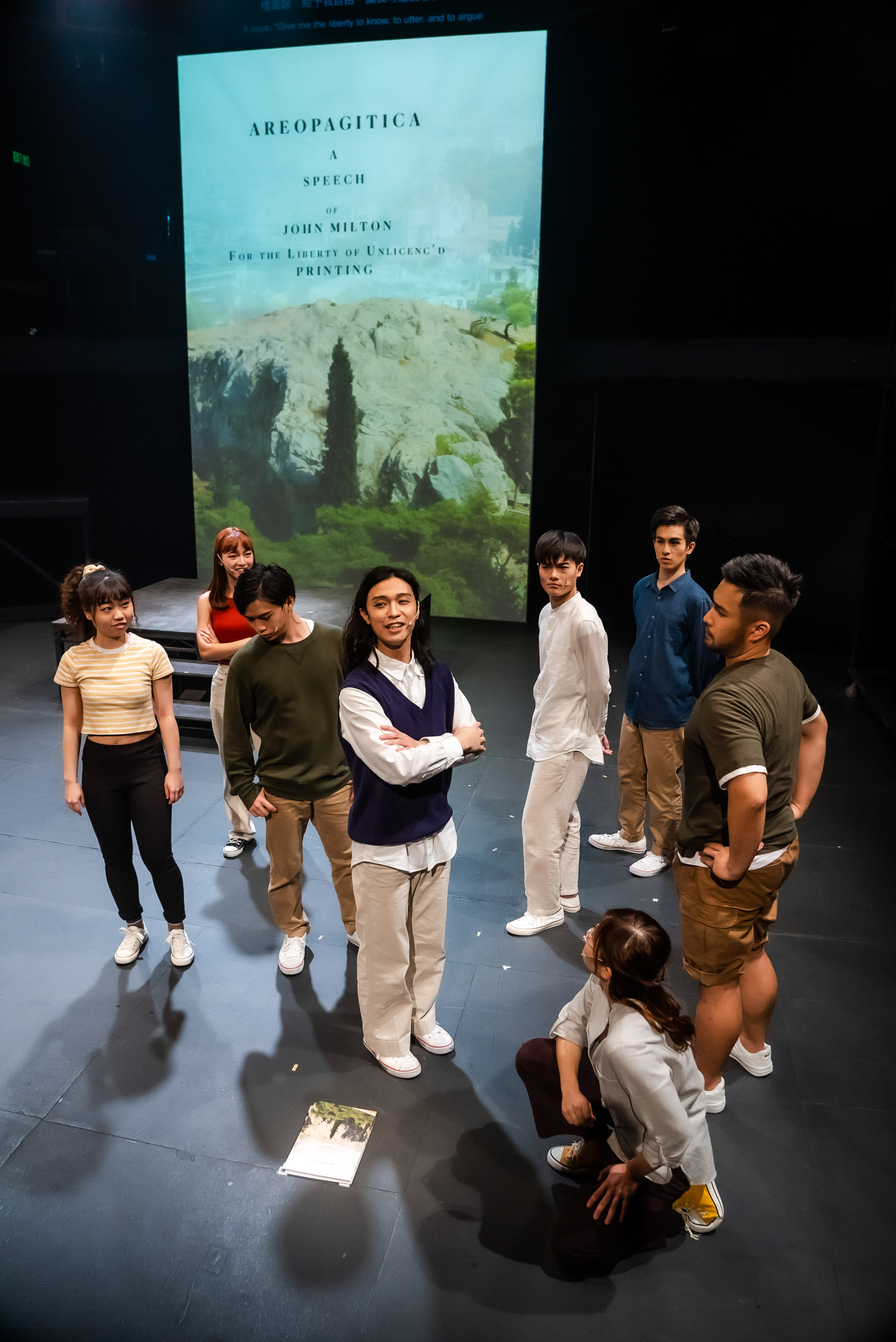 一幅充滿活力的香港當代劇團《天真與世故之歌》舞台表演場景，九位演員站在簡潔而燈光明亮的舞台上。背景的大型投影螢幕上顯示著「AREOPAGITICA A SPEECH OF JOHN MILTON FOR THE LIBERTY OF UNSLICENC'D PRINTING」的文字，並配以岩石與翠綠景觀的圖像。在群體中央，麥晧鈿（可能飾演密爾頓）自信地交叉雙臂站立，身穿白襯衫和深藍色V領背心，臉上帶著淺淺的微笑。在他左側，冼樂欣（可能飾演夏娃或天使）穿著條紋露臍上衣和黑色緊身褲，表情投入。附近，莫珏邦（可能飾演米迦勒）身穿橄欖綠毛衣，目光下垂，而另一位穿紅色上衣的女演員則站在他身後。在麥晧鈿右側，張卓行（可能飾演天使）身穿寬鬆白襯衫站立，在他身後，趙汝陽（可能飾演瑪門）身穿深藍色襯衫和米色褲子，目光望向左側。最右邊，一位身穿深綠色T恤和棕色短褲的男演員雙手叉腰站立。前景中，李安生或劉嘉盈（可能飾演摩洛或彼列）蹲在深色舞台地板上，抬頭仰望，她面前還放著一本打開的書或劇本。強烈的舞台燈光投射出清晰的演員剪影，突顯了這場富於感染力的舞台表演中，演員們的存在感與專注。