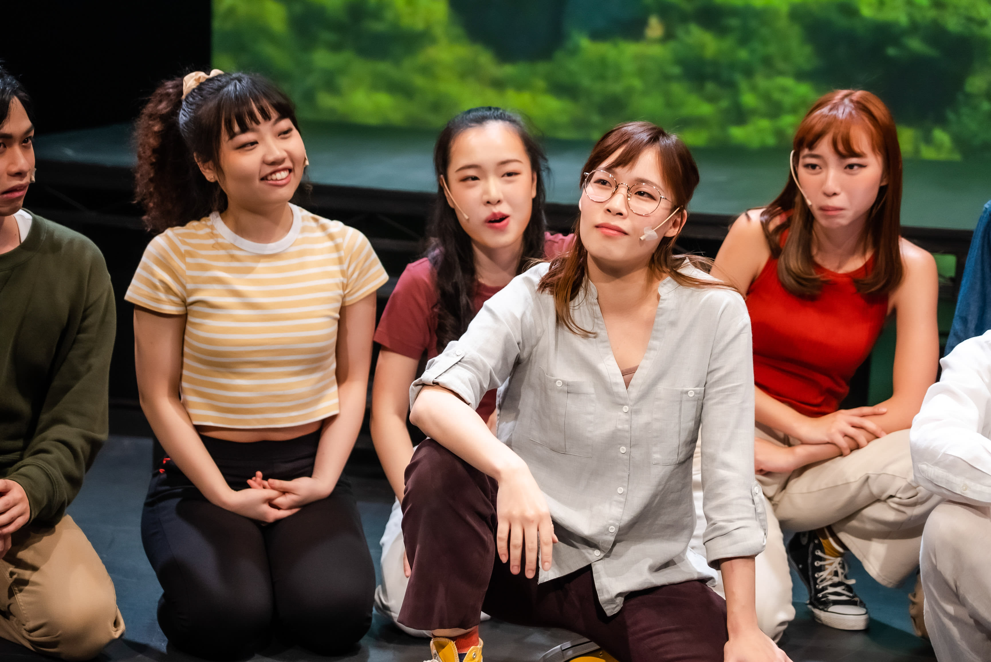 Six actors from the contemporary Hong Kong theatre production 'Songs of Innocence and Experience' are seated and kneeling on a dark stage against a blurry green background. From left, Chew Yu-yeung (Mammon) looks thoughtful. Sin Lok-yan (Eve / Angel) in a striped top smiles broadly, clasping her hands. Behind her, Sum Cheuk-yiu (Abdiel) in red, appears to be singing or speaking. In the foreground center, Lau Ka-ying (Belial) wears glasses and a light grey shirt, looking contemplatively upwards with a visible microphone on her cheek. To her right, Lee On-sang (Moloch) in a red tank top also has a mic and a pensive expression. Another actor is partially visible on the far right. The diverse cast is engaged in a reflective moment, characteristic of a live stage performance exploring themes of youth and personal growth. So... 'What is growing up?'