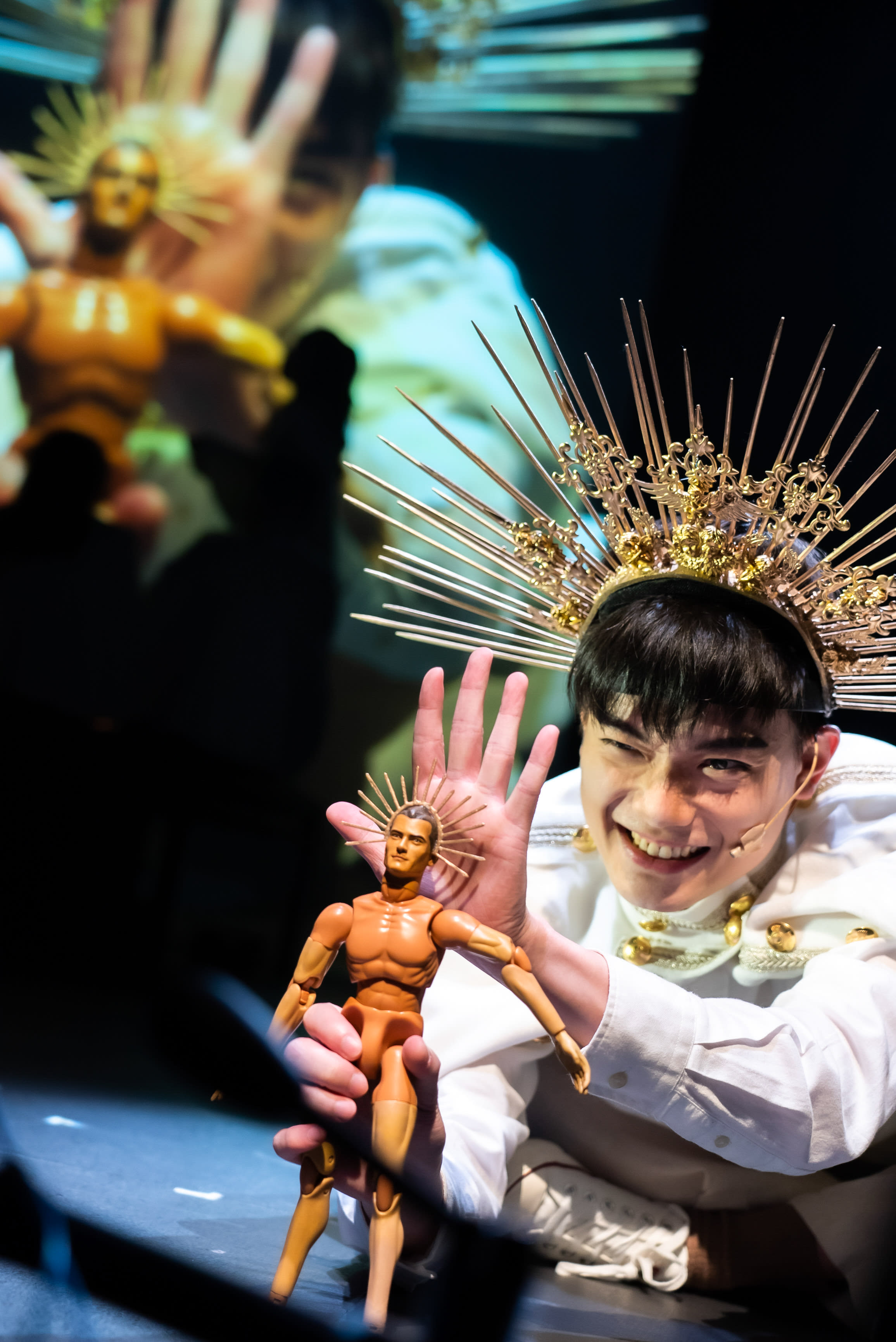 In a captivating scene from Rooftop Productions' contemporary HK theatre show, Songs of Innocence and Experience, actor Lo Man-chak, portraying 'Himself / God,' grins playfully at the audience, winking. He wears a majestic golden sunburst crown and a white theatrical costume. With his left hand, he holds up a muscular action figure, similarly adorned with a halo, while his right hand is raised behind it in a gesturing pose. The dramatic stage lighting emphasizes his joyful expression and the intricate details of the props against a dark, softly projected background that mirrors the halo motif, encapsulating the vibrant energy of this unique stage performance. Today, I’m going to introduce my newest creation - my son!