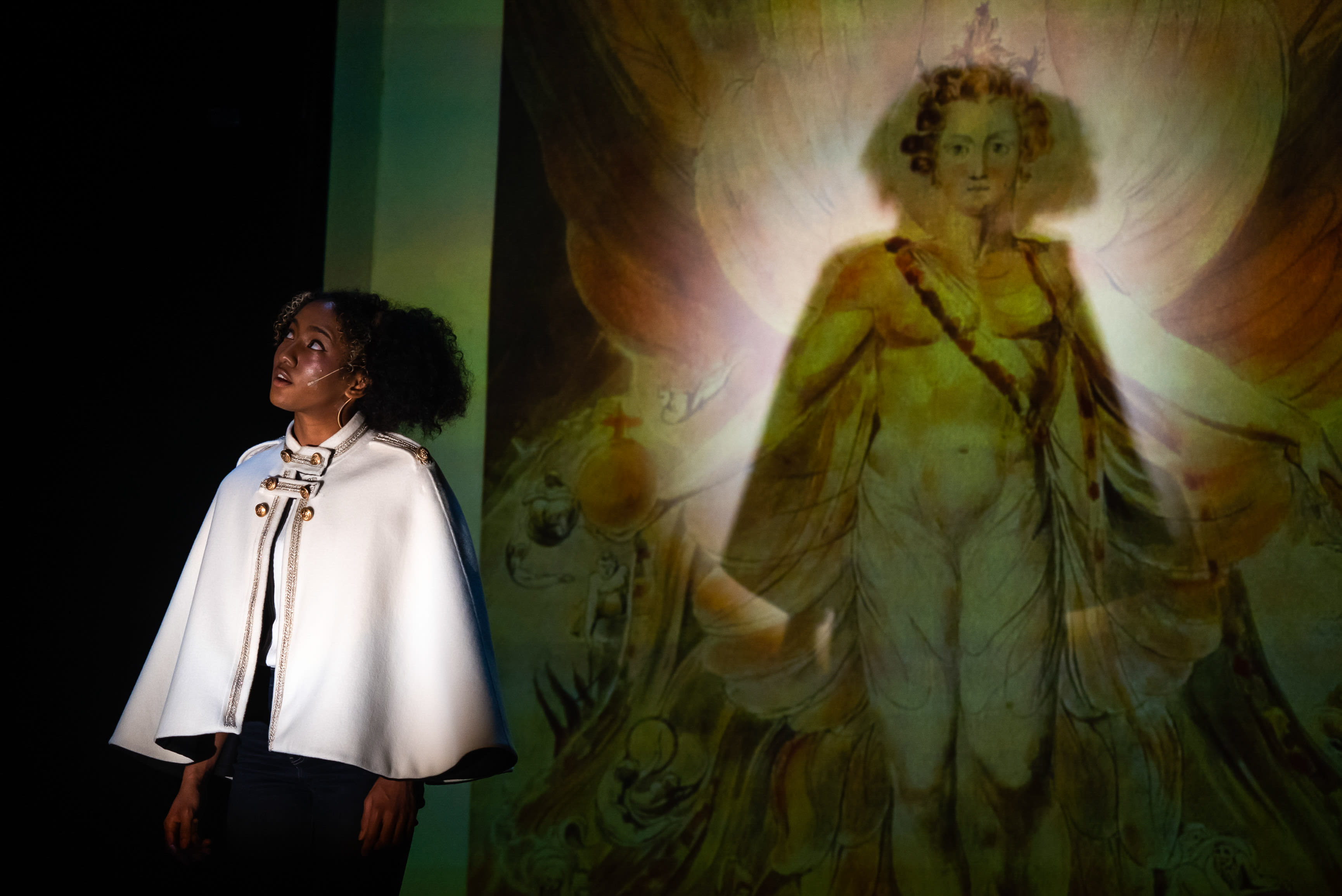 Caroline Chan, portraying 'Herself' or Satan (Lucifer) in Rooftop Productions' contemporary HK theatre production 'Songs of Innocence and Experience,' stands under a spotlight on the left, looking upwards and to the right with an expression of awe or profound questioning. She wears a striking white cape adorned with gold and black military-style accents, over a dark outfit, with a small microphone visible. To her right, a large, ethereal projection fills the stage backdrop, depicting a majestic, crowned, androgynous figure with an antique aesthetic, emanating light and surrounded by a powerful aura, reminiscent of a classical painting. This dramatic stage performance creates a powerful visual contrast between the actor and the projected divine image. God, are you really all-powerful?