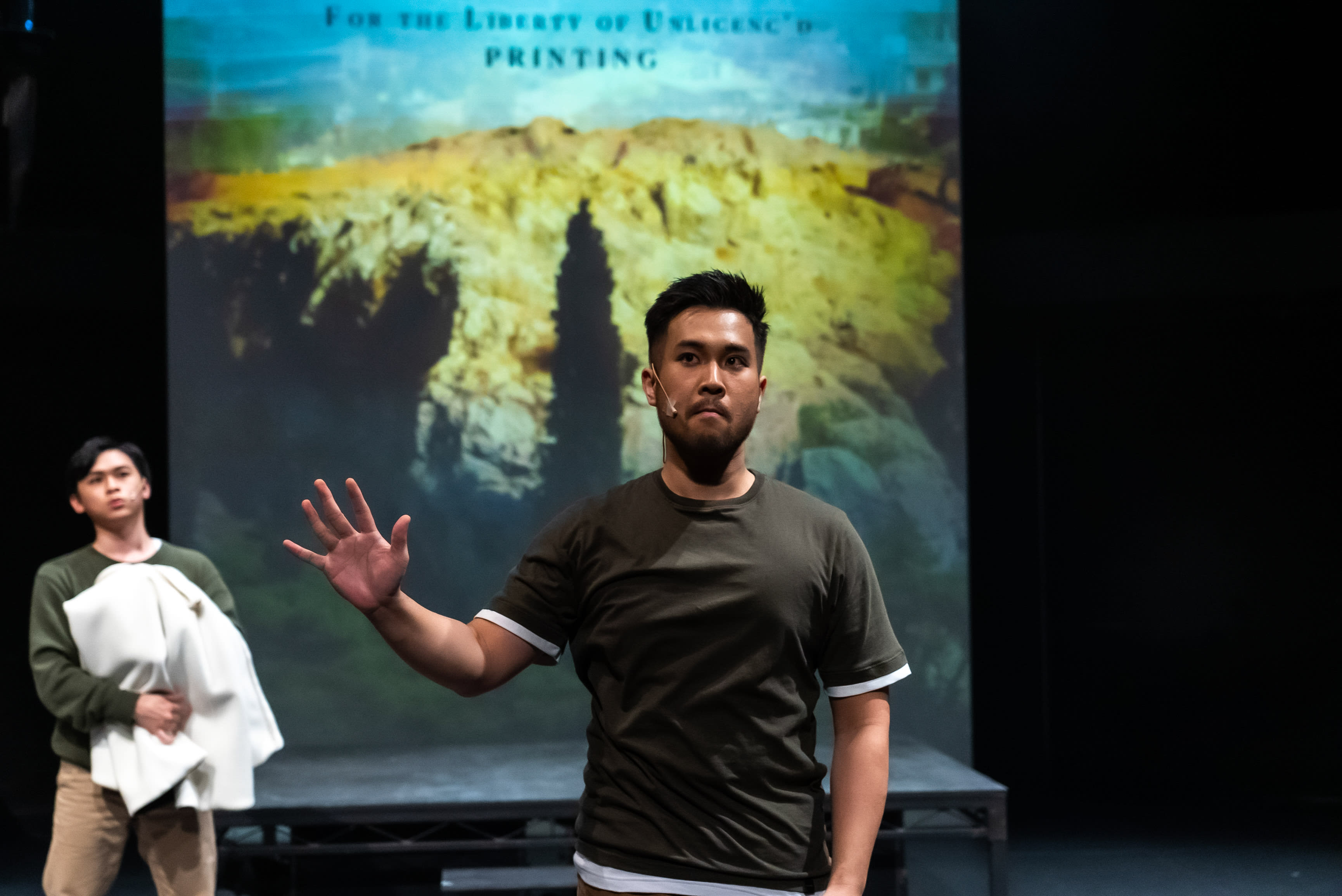 在天台製作《天真與世故之歌》的昏暗舞台上，演員莫珏邦（飾演米迦勒）身穿深綠色T恤，配有白色袖口和下擺，站在舞台中央偏右，舉起右手，掌心向左，神情嚴肅，留著短鬚，佩戴著隱形耳機麥克風。舞台左後方，演員趙汝陽（飾演瑪門）身穿綠色長袖上衣和卡其褲，手抱一件折疊的白色衣物，目光專注地望向右上方。兩人身後是一個巨大的投影屏幕，展示著一片黃褐色崎嶇山脈和藍天景觀，屏幕頂部可見「FOR THE LIBERTY OF UNLICENC'D PRINTING」字樣，為這場香港當代舞台劇營造出強烈的戲劇氛圍。叛變... 我不會的
