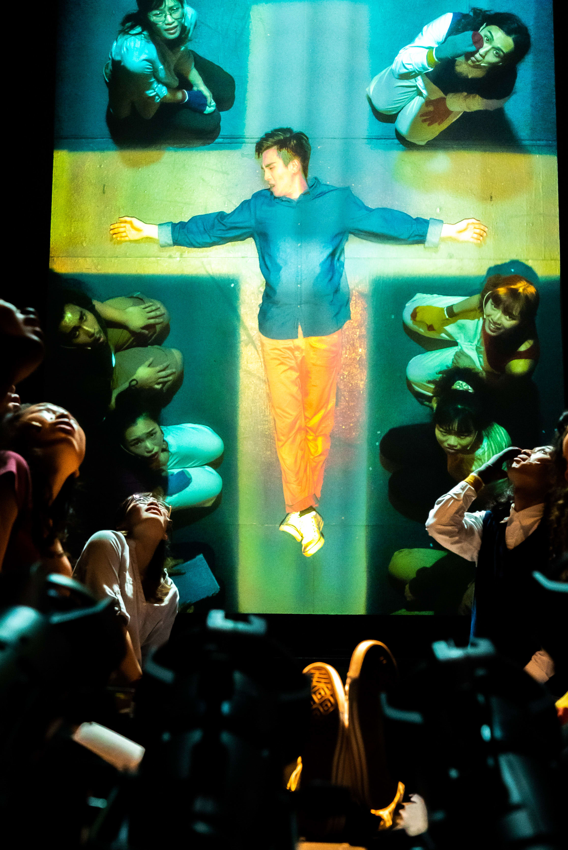 In this evocative scene from Rooftop Productions' contemporary Hong Kong theatre piece 'Songs of Innocence and Experience', a central male figure, portrayed by Mok Kok-pong as Michael, is suspended horizontally in a striking cruciform pose against a floor-lit cross. Surrounded by fellow performers including Sin Lok-yan as Eve and other angels and fallen spirits, the dramatic lighting highlights the intensity of this avant-garde stage performance. One performer cries out, 'Sorry, Jesus, please, have pity on me!'