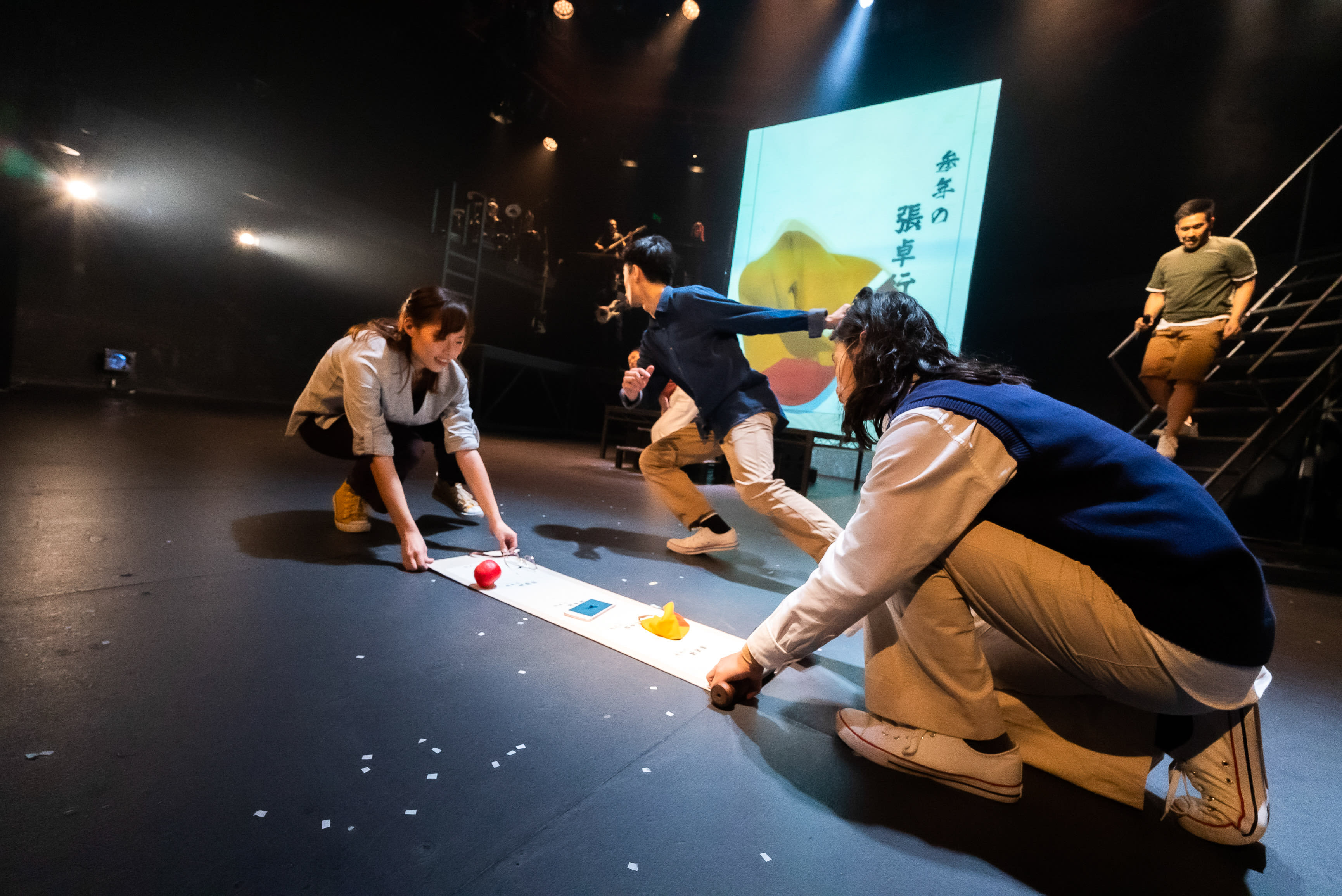 在一個昏暗的舞台上，四位演員正在一場香港當代劇場演出中進行互動。劉嘉盈（飾演彼列）在左側蹲下，小心翼翼地將一個紅色球體放在地上長長的白色條帶上。在她的右邊，張卓行（飾演天使）也蹲著，觀察著條帶上的黃色物體。麥晧鈿（飾演密爾頓／別西卜）在舞台中央呈現衝刺狀，充滿活力地奔向活動。背景中，莫珏邦（飾演米迦勒）在金屬樓梯上可見，正在觀看。後方的大螢幕上投射出劇團Rooftop Productions（天台製作）的劇目《天真與世故之歌》的中文標題及抽象的黃色和紅色圖像。舞台上散落著白色紙屑，聚光燈照亮了這些動態的場景，展現了這場香港當代舞台表演的活力。如同對這場熱烈互動的邀請，表演者們體現著劇中台詞的精神：「來，戰鬥吧！」
