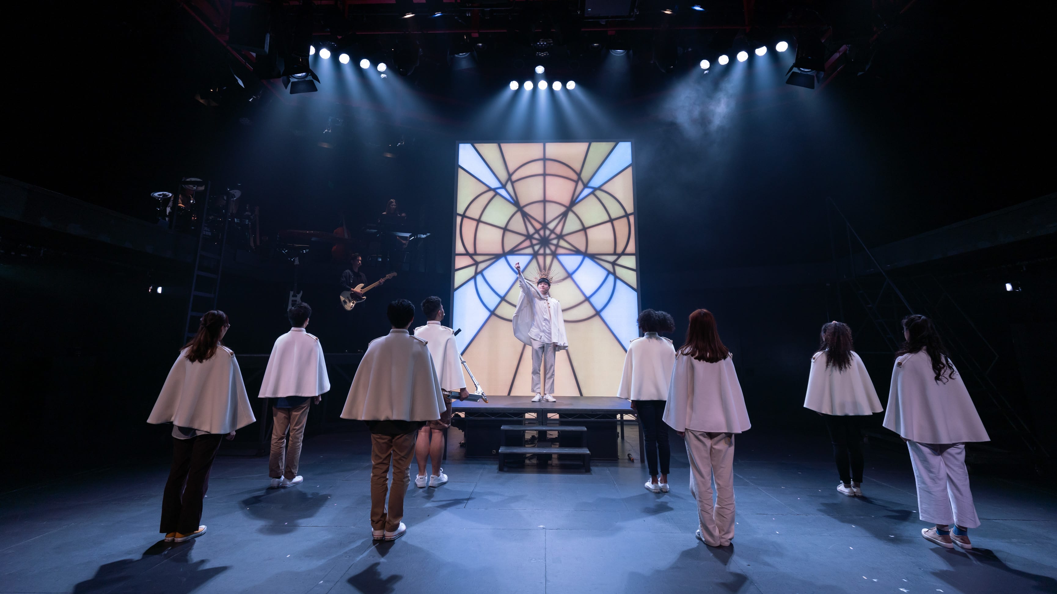 A dramatic stage scene from Rooftop Productions' contemporary theatre performance, 'Songs of Innocence and Experience' (《天真與世故之歌》). At the center, Lo Man-chak, portraying God, stands elevated on a platform in front of a striking, illuminated geometric backdrop that resembles a stained-glass window. Below, a group of cast members—including performers representing entities like Abdiel, Belial, Moloch, Mammon, Michael, Satan, and Eve—stand with their backs to the audience, wearing white capes, as if listening to a divine pronouncement. The atmospheric lighting creates a cinematic quality typical of professional Hong Kong stage productions. Lo Man-chak, as God, commands the space: 'Hear all ye Angels, Thrones, Dominions.'