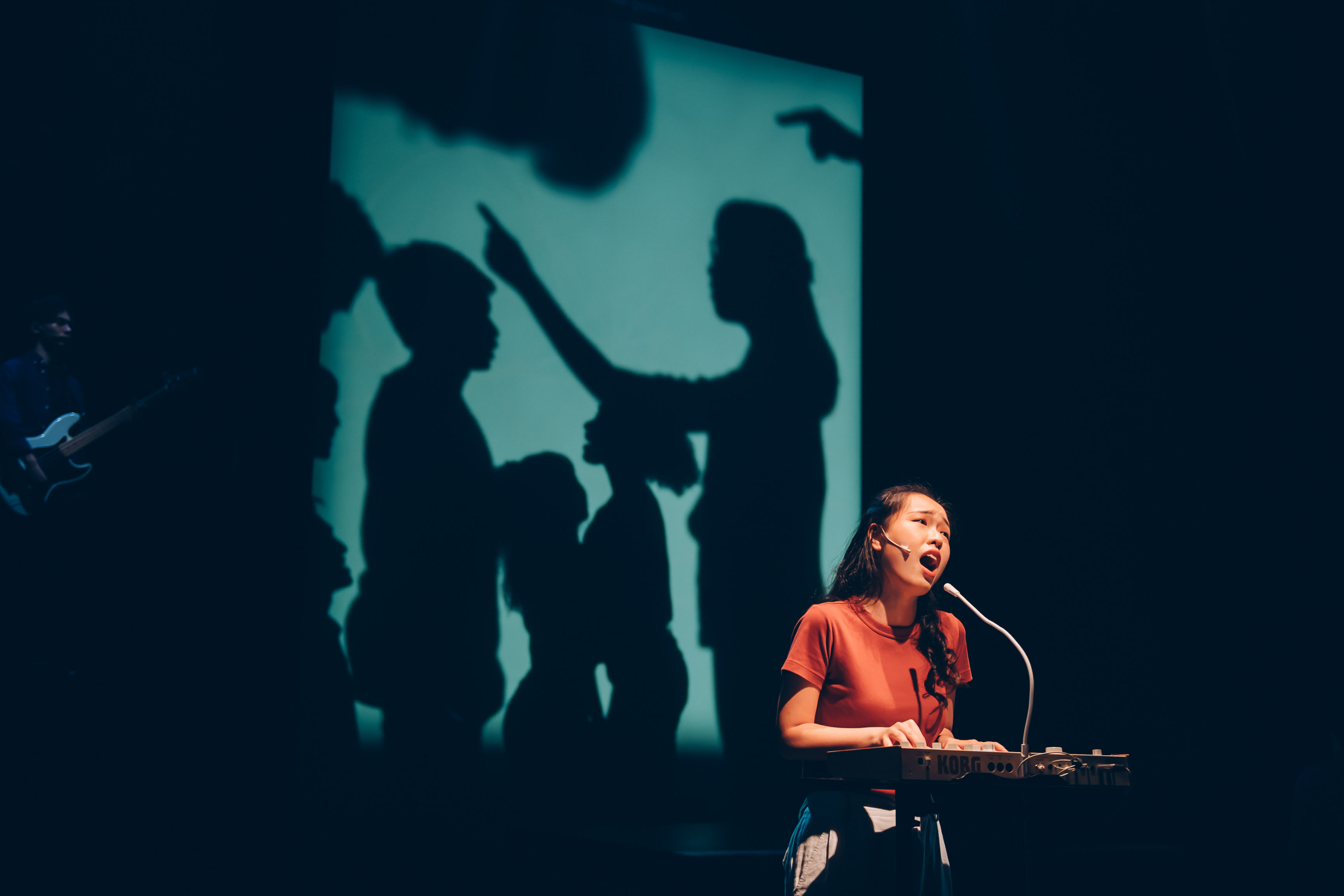 沈卓瑤在香港當代劇場作品《天真與世故之歌》中現場演出，她身穿鐵鏽色T恤，在電子鍵盤前深情地對著麥克風歌唱。舞台昏暗，她被明亮的聚光燈照亮，身後的大型青藍色屏幕上，投射出數個引人注目的剪影，其中一人手指著前方，暗示著戲劇性的互動或衝突。在後方昏暗的角落，一名樂手正彈奏電吉他，為這場多媒體舞台表演增添層次。 「令媛已長大了」