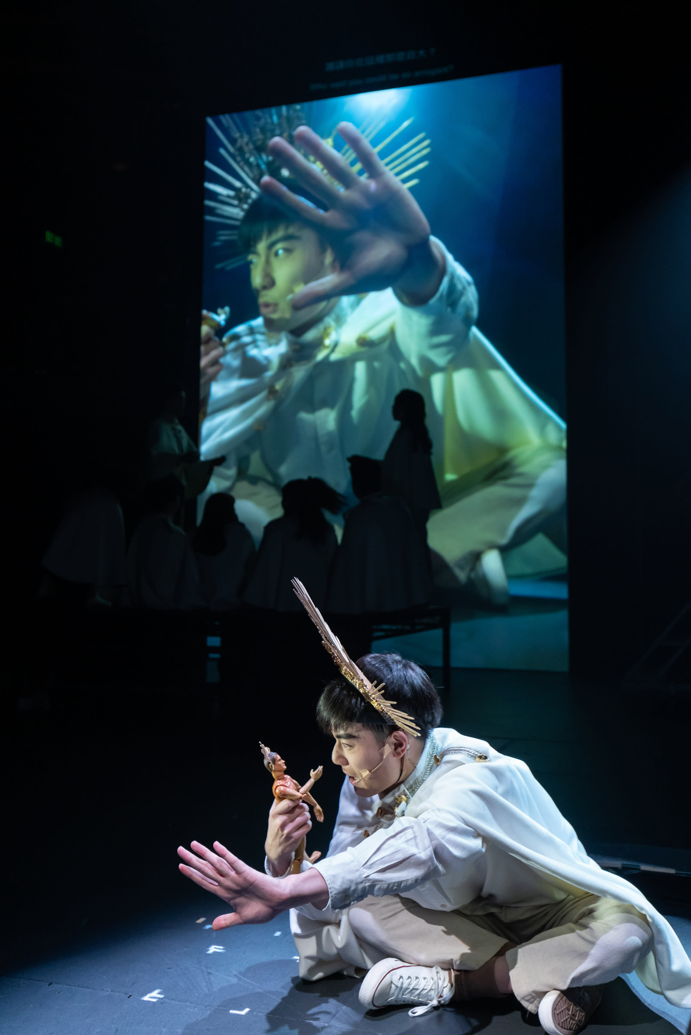 在天台製作（Rooftop Productions）的當代劇場作品《天真與世故之歌》中，演員羅文澤身穿白色服裝，頭戴金屬頭飾，坐在昏暗的舞台上。他手中握著一個玩偶，表情生動地與其互動，動作與背景大螢幕上放映的巨型投影相互呼應。這種真人表演與影像投射交織的藝術手法，展現了香港當代劇場獨特的實驗性舞台美學。他在表演中對玩偶說道：「不肖子！誰讓你在這裡那麼自大？」