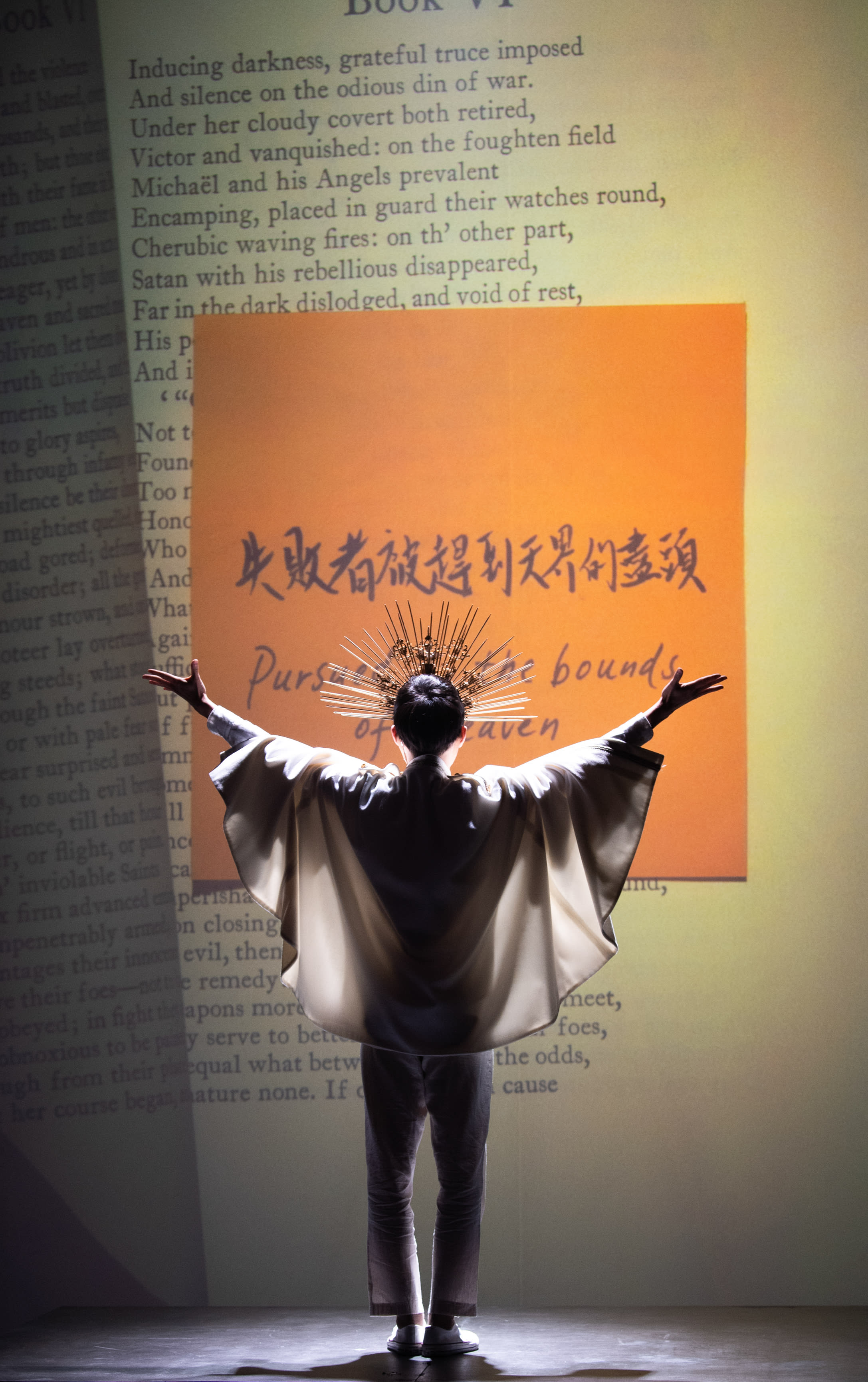 A dramatic stage shot from Rooftop Productions’ contemporary theatre piece 'Songs of Innocence and Experience'. The performer Lo Man-chak, portraying God, stands with his back to the audience against a large, golden-orange backdrop layered with English text from Milton’s Paradise Lost and bold Chinese calligraphy that reads 'The losers are driven to the edge of heaven'. He wears an elaborate headpiece resembling a golden sunburst and a flowing white garment with wide sleeves, his arms outstretched in a powerful, celestial gesture. The lighting focuses sharply on his silhouette, emphasizing the scale and spiritual weight of the performance. As he stands at the horizon of his own creation, Lo Man-chak delivers the line: 'When I am alone, I dream, staring at the horizon, but words fail.'