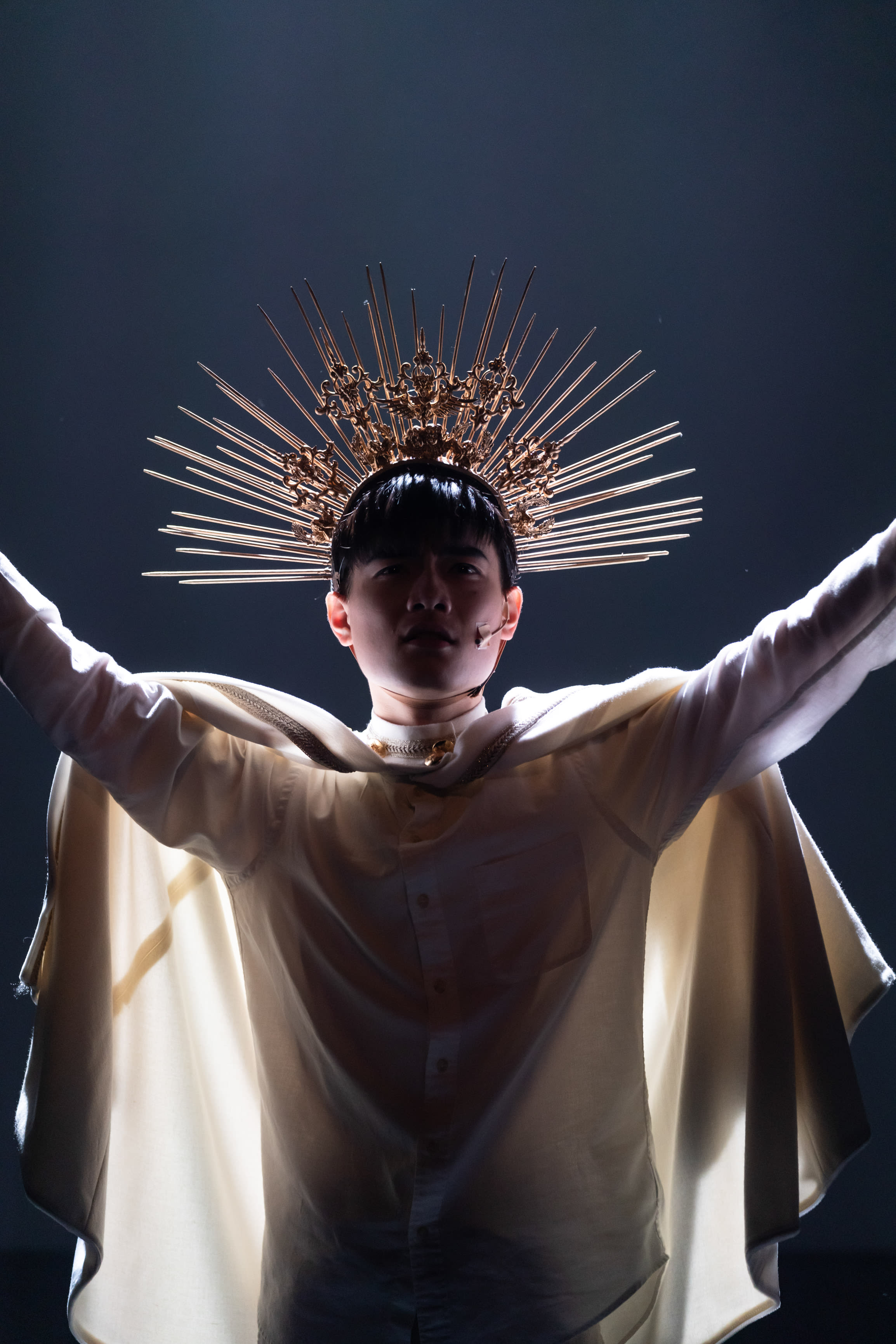 Lo Man-chak stands center stage in a dramatic, high-contrast spotlight, portraying God in Rooftop Productions' contemporary theatre piece, Songs of Innocence and Experience. He wears a radiant gold sunburst crown and a flowing white cape over a modern shirt, arms outstretched in a divine, solemn posture against a dark, moody background. As he embodies the role, he delivers the haunting line: "Yes, I know that there is no light in a room when the sun is missing."