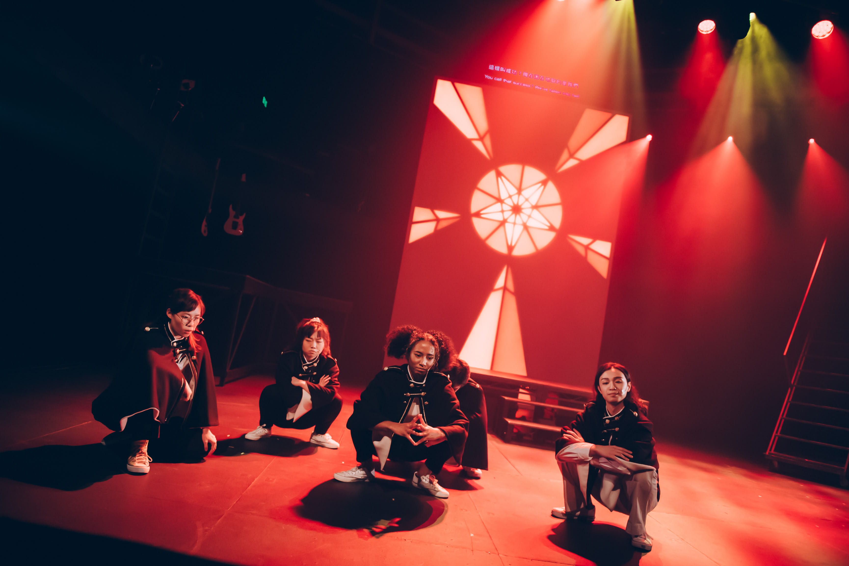 Four actors from Rooftop Productions perform on a dark, red-lit stage in the contemporary Hong Kong theatre production 'Songs of Innocence and Experience' (《天真與世故之歌》). Positioned in a dramatic crouch under a glowing, geometric stained-glass backdrop, the cast members—representing figures such as Satan, Moloch, Belial, and Mammon—exude a tense, powerful energy under the intense stage lighting. As they reflect on their journey, the ensemble captures the essence of the performance, echoing: "Although the results weren't ideal, our challenge was a success."