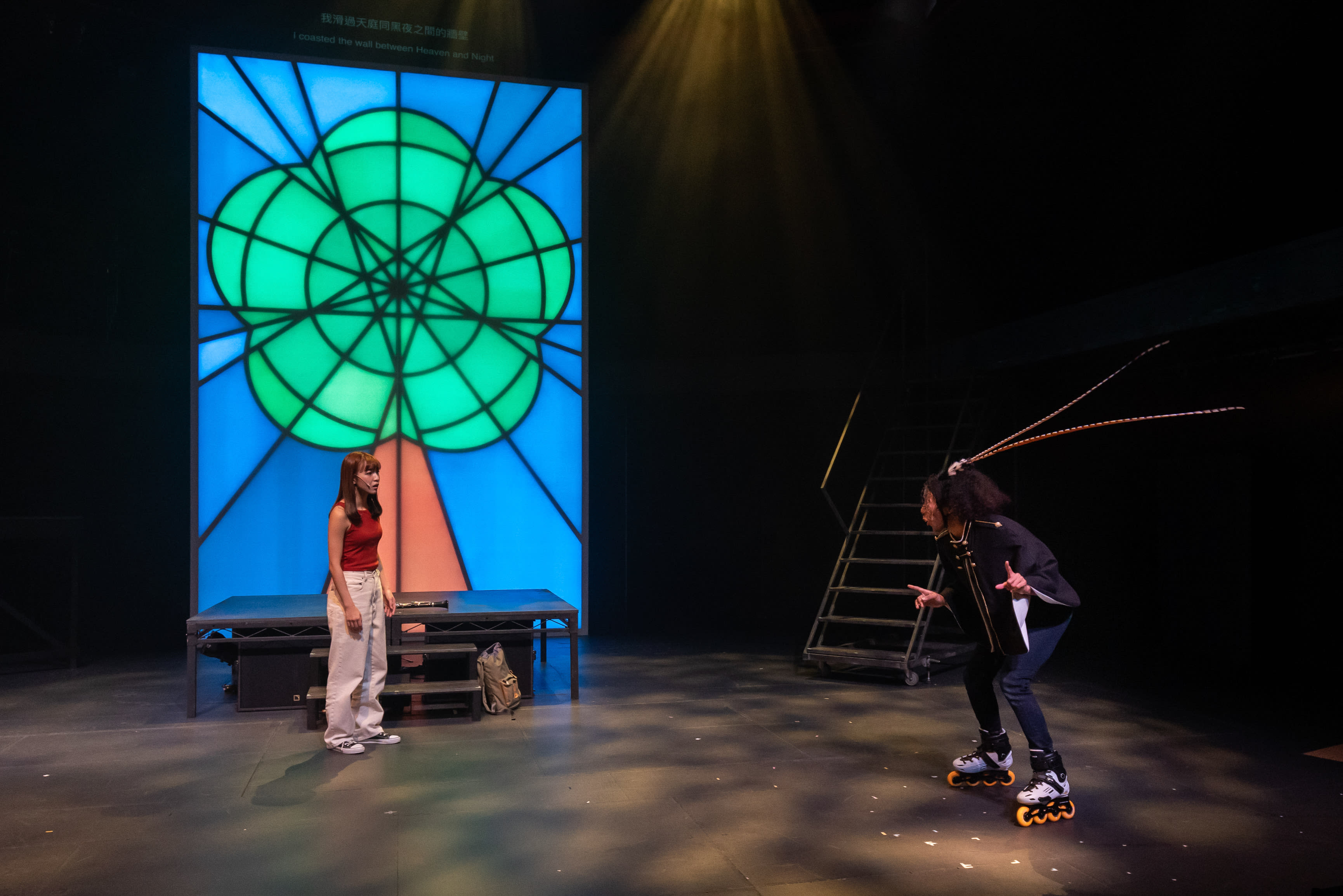 天台製作（Rooftop Productions）當代劇場演出《天真與世故之歌》的戲劇一幕：演員冼樂欣身穿紅色上衣配米色長褲，站在舞台左側；右側的演員陳琬瑜則穿著黑色服飾並腳踏直排滑輪鞋，身體前傾，展現出充滿張力的姿態，頭上戴著模仿觸角或尾巴的舞台道具。舞台燈光聚焦於兩人，背景是一個巨大的幾飾風格綠色花朵造型背光裝置。飾演夏娃的演員說：「我⋯又沒有想過要走。」
