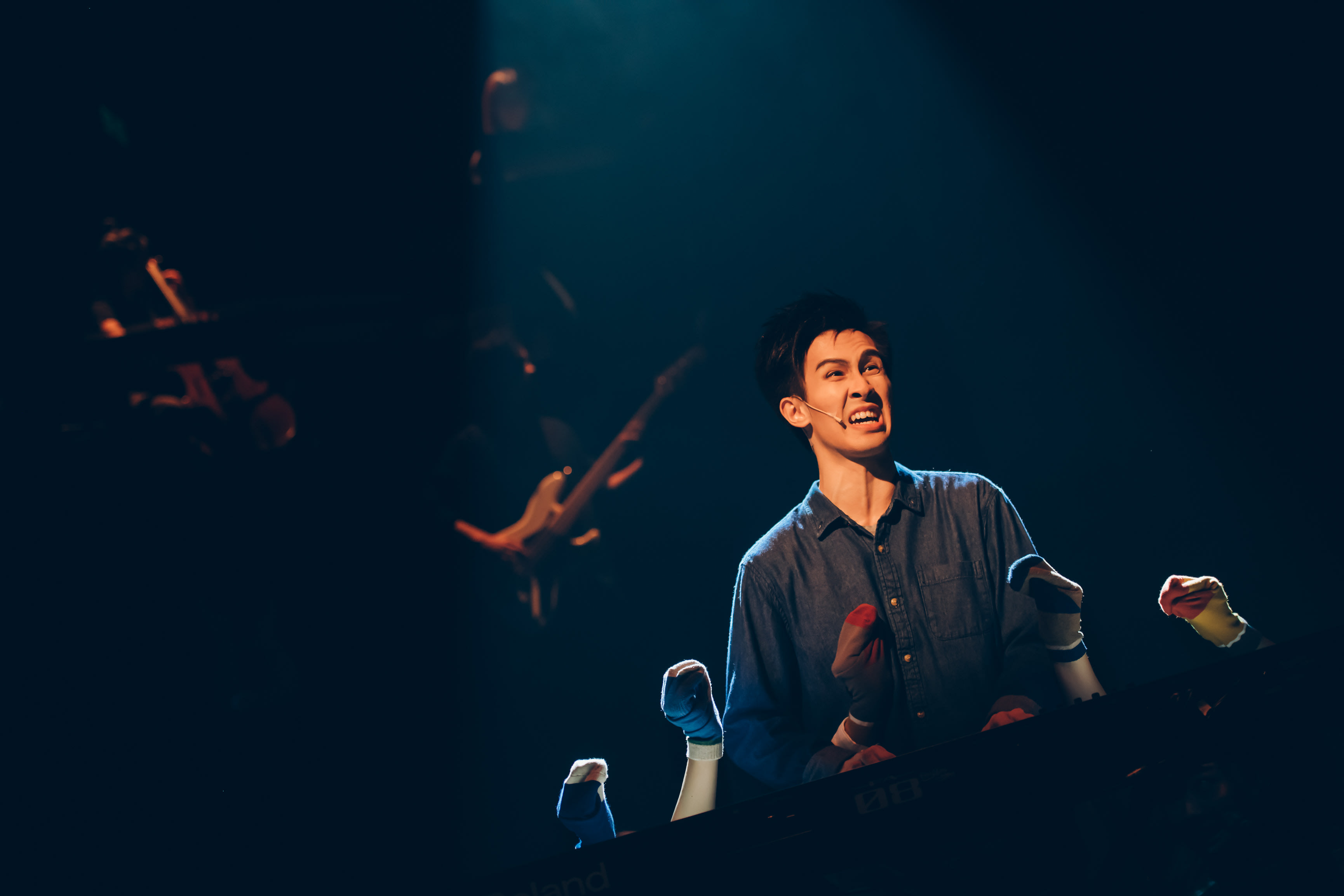 In a dramatic spotlight during a Rooftop Productions (天台製作) contemporary theatre performance of 'Songs of Innocence and Experience', actor Cheung Cheuk-hang, playing the role of himself/Angel, performs with an intense, pained expression. He is positioned behind a keyboard, surrounded by multiple disembodied sock puppets arranged as if playing instruments, set against a dark stage atmosphere. As he sings with raw emotion, he poses the poignant question: "Have you sacrificed anything to help others?"