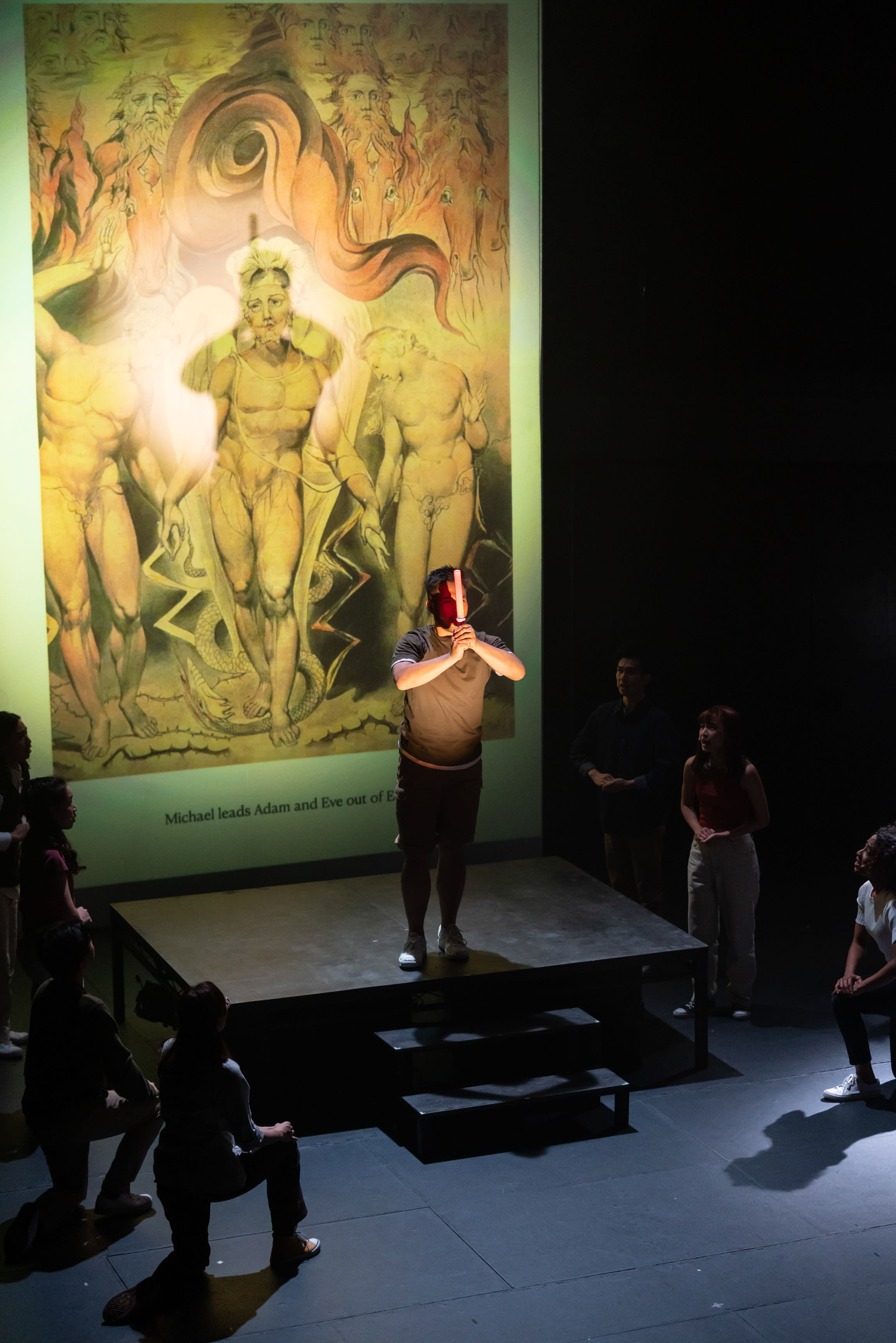 A powerful scene from Rooftop Productions' contemporary HK theatre performance, "Songs of Innocence and Experience," features actor Mok Kok-pong portraying Michael on a raised stage. He stands center, illuminated by a bright spotlight, holding a red rectangular prop that obscures his face. Behind him, a large projection displays William Blake's artwork depicting Michael leading Adam and Eve out of Eden, reinforcing the play's themes of innocence and experience. Other cast members are dimly visible around the dark stage platforms, observing this dramatic moment. Mok Kok-pong (as Michael) proclaims, "My name is Michael too."