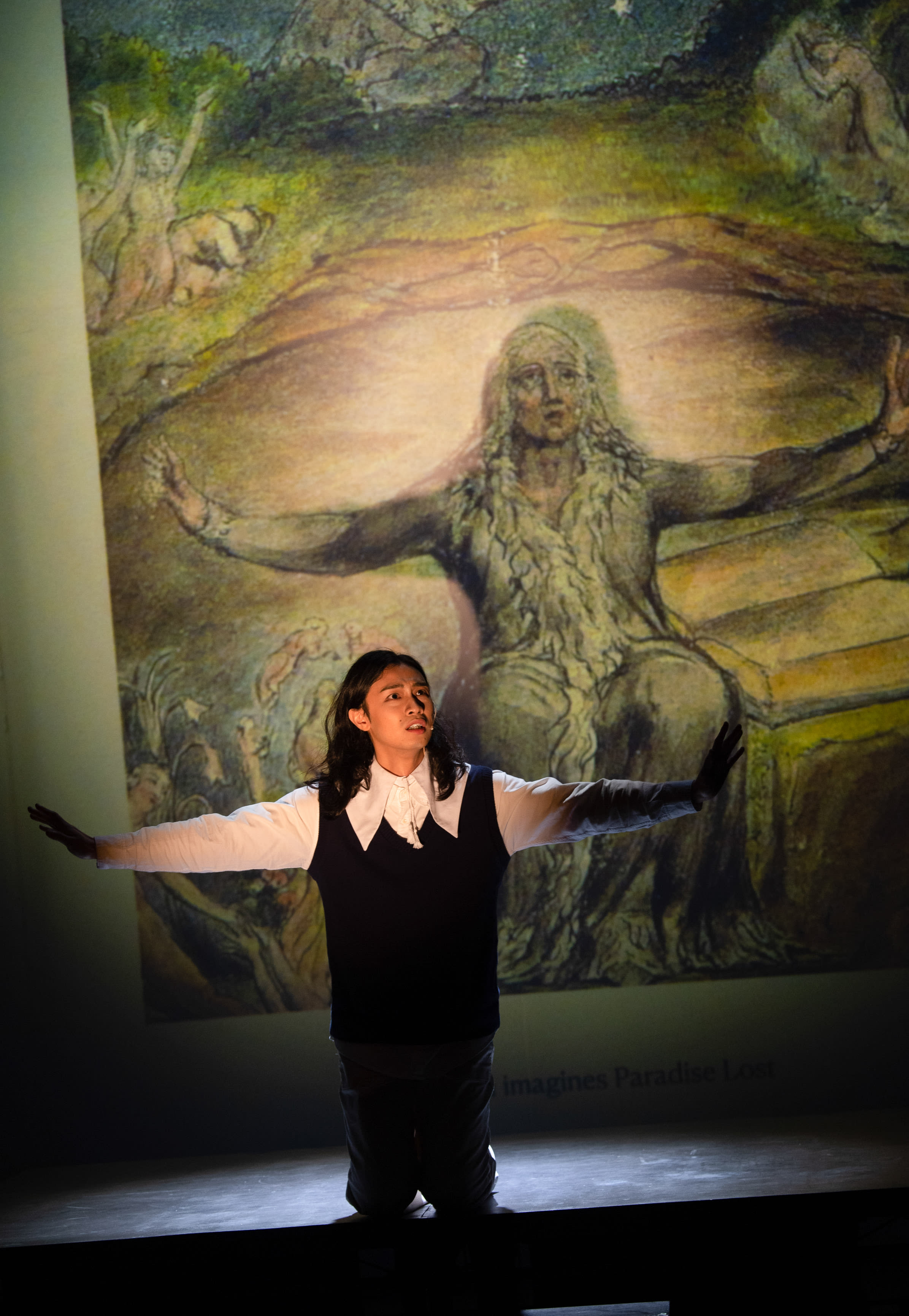 In a dramatic scene from Rooftop Productions' contemporary theatre piece 'Songs of Innocence and Experience', actor Mak Ho-tin kneels on a dimly lit stage, his arms outstretched in a mirror image of the large, expressive William Blake illustration projected behind him. The atmospheric lighting emphasizes his emotive performance as he embodies Milton, declaring, 'History will remember me as a loser.'