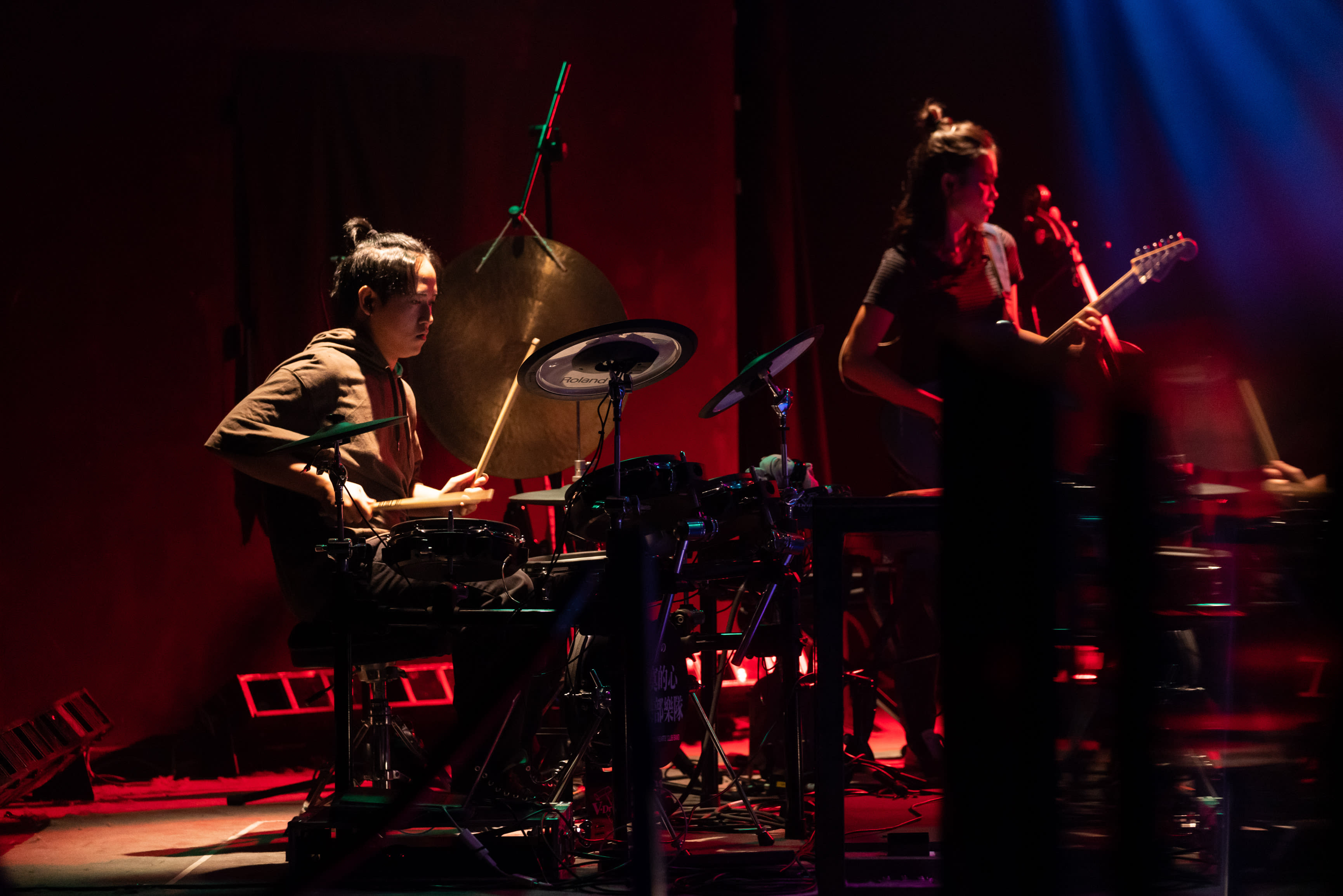 In a dramatic scene from Rooftop Productions' contemporary theatre performance, 'Songs of Innocence and Experience,' Sung Kwok-tung is seen focused on playing electronic drums and a large gong under moody, deep red stage lighting, while Chin Wing-kar plays an electric guitar in the background. This atmospheric production explores complex themes of musical storytelling in Hong Kong theatre. As the tension mounts, a performer sings, "Don't cry out loud."