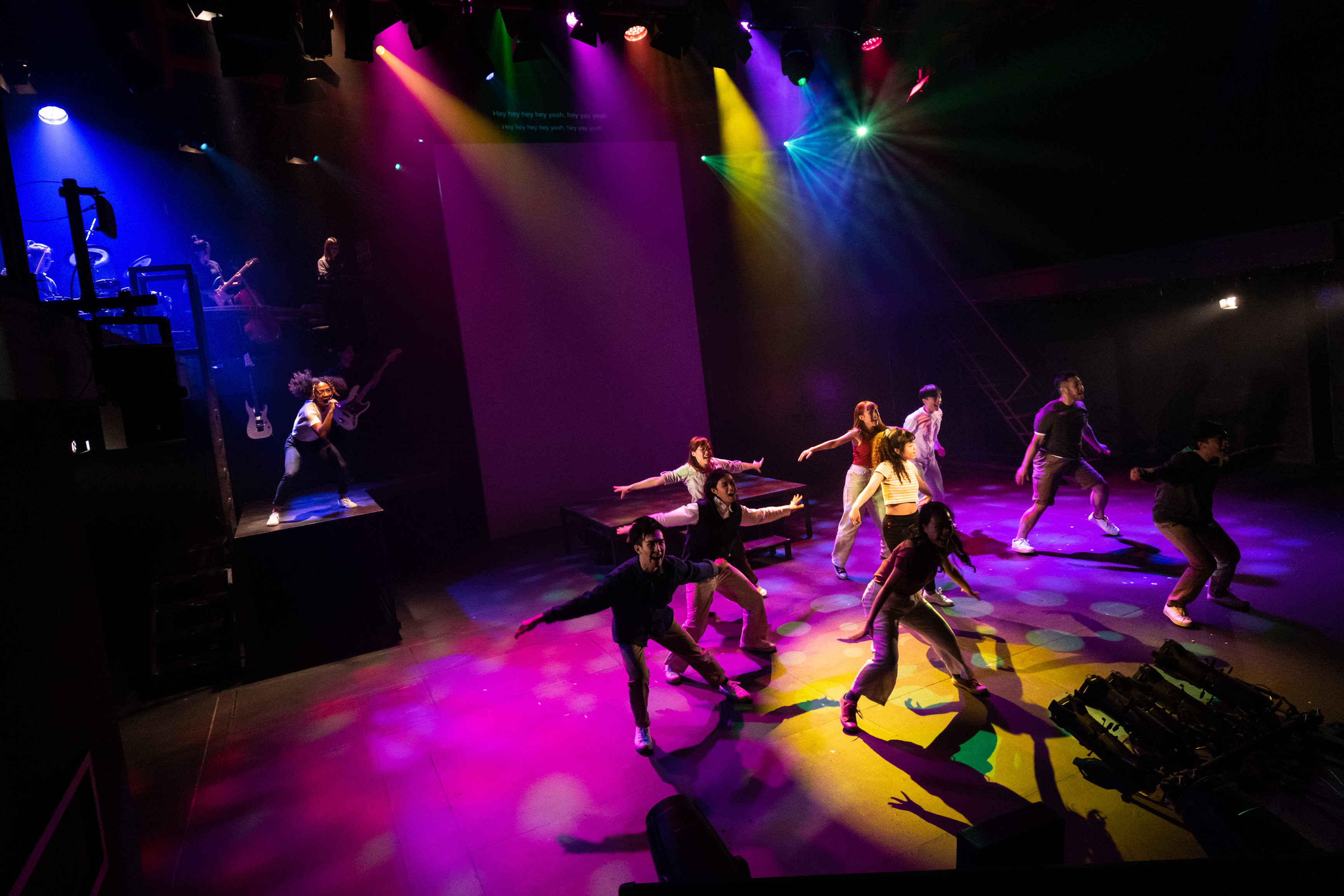 A high-energy moment from Rooftop Productions' contemporary HK theatre performance, "Songs of Innocence and Experience," unfolds on a stage illuminated by a kaleidoscope of purple, blue, yellow, green, and red spotlights. A large purple backdrop complements the vibrant floor, which reflects the dramatic lighting. A diverse ensemble of performers, including Caroline Chan as Satan (Lucifer) in a powerful lunge in the foreground, and Sum Cheuk-yiu as Abdiel and Lee On-sang as Moloch among others, are captured in dynamic, expressive movements across the stage. On a raised platform to the left, Wu Lui-fung, the Band Leader, plays an electric guitar, providing live musical accompaniment for this compelling stage performance. The scene bursts with theatrical intensity, capturing a moment of palpable drama. A character asks, "What's going on?"
