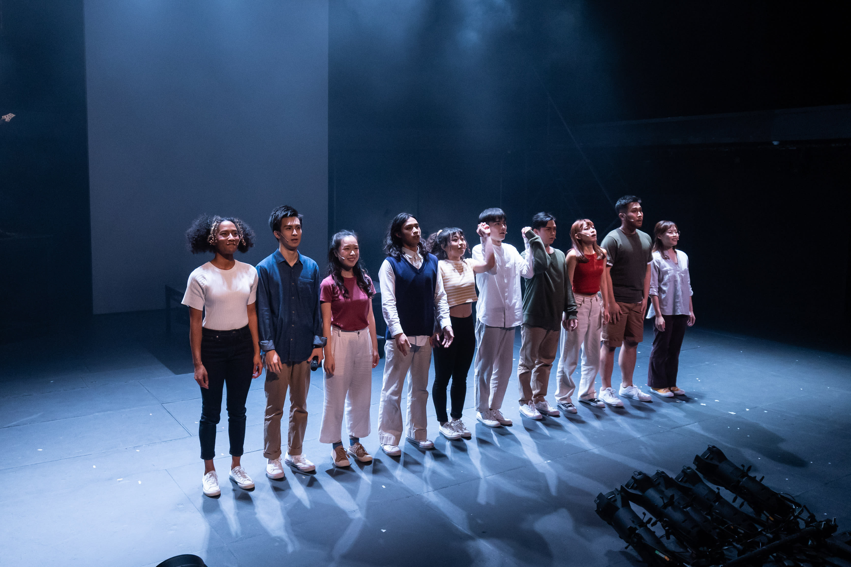 A group of twelve performers, including Sum Cheuk-yiu, Cheung Cheuk-hang, Lau Ka-ying, Chew Yu-yeung, Sin Lok-yan, Mak Ho-tin, Caroline Chan, Mok Kok-pong, Lo Man-chak, and Lee On-sang, stand in a line across a dark stage, illuminated by a spotlight from above. Their faces are turned upwards and slightly to the right, with expressions ranging from contemplative to questioning, embodying the themes of Rooftop Productions' contemporary Hong Kong theatre piece, *Songs of Innocence and Experience*. The bare stage and dramatic lighting emphasize the actors' presence in this powerful stage performance. One of the performers appears to be speaking the line, "I said hey, what's going on?"