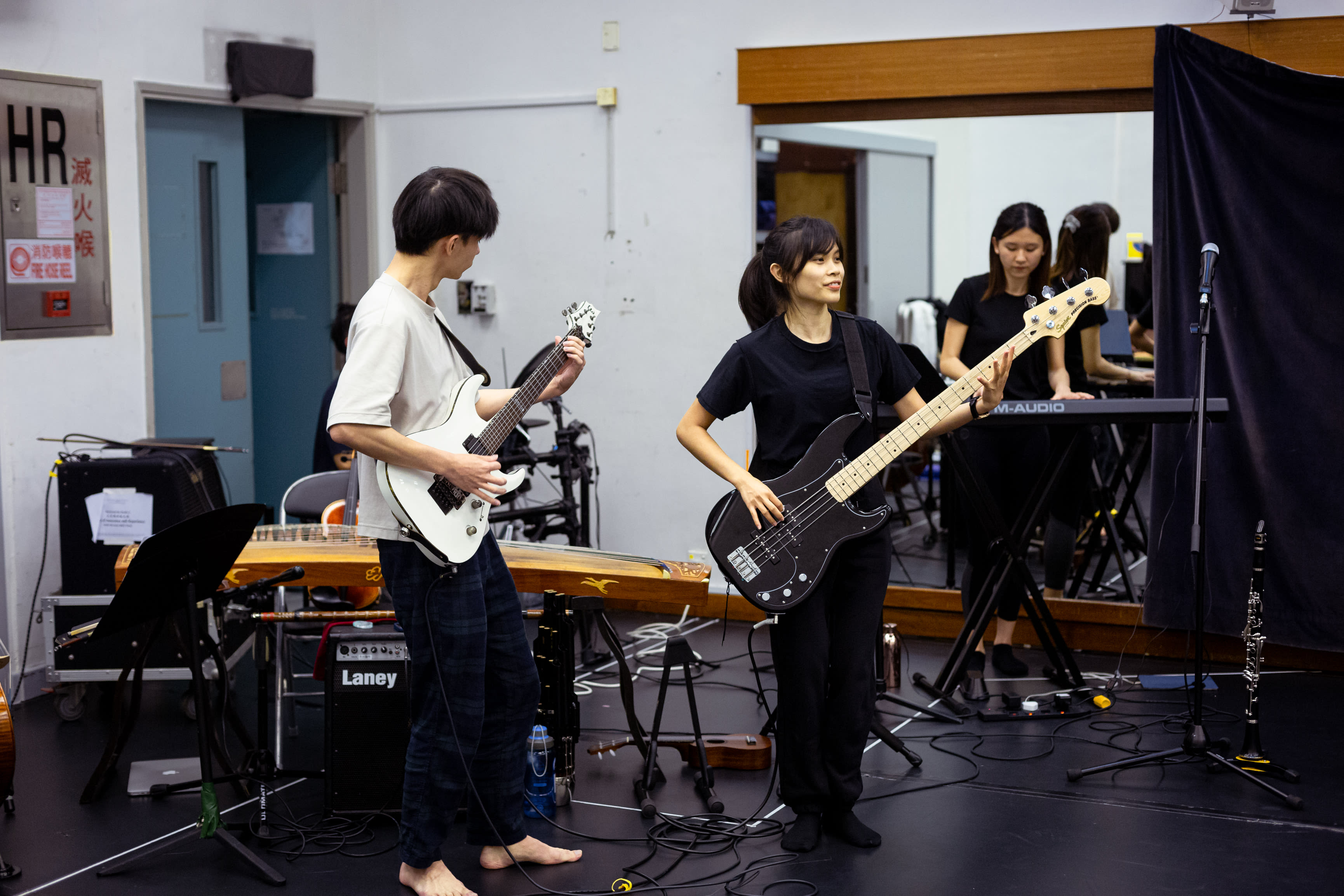 In this rehearsal for Rooftop Productions' contemporary theatre piece 'Songs of Innocence and Experience', band leader Wu Lui-fung stands on the left playing a white electric guitar, while musician Chin Wing-kar stands on the right, focused as she plays a black electric bass. In the background, musician Tsang Yuen-lam is captured in a large wall mirror practicing on a keyboard. The room is a functional studio space, equipped with various musical instruments including a guzheng and amplifier. Songs of Innocence and Experience Rehearsal.